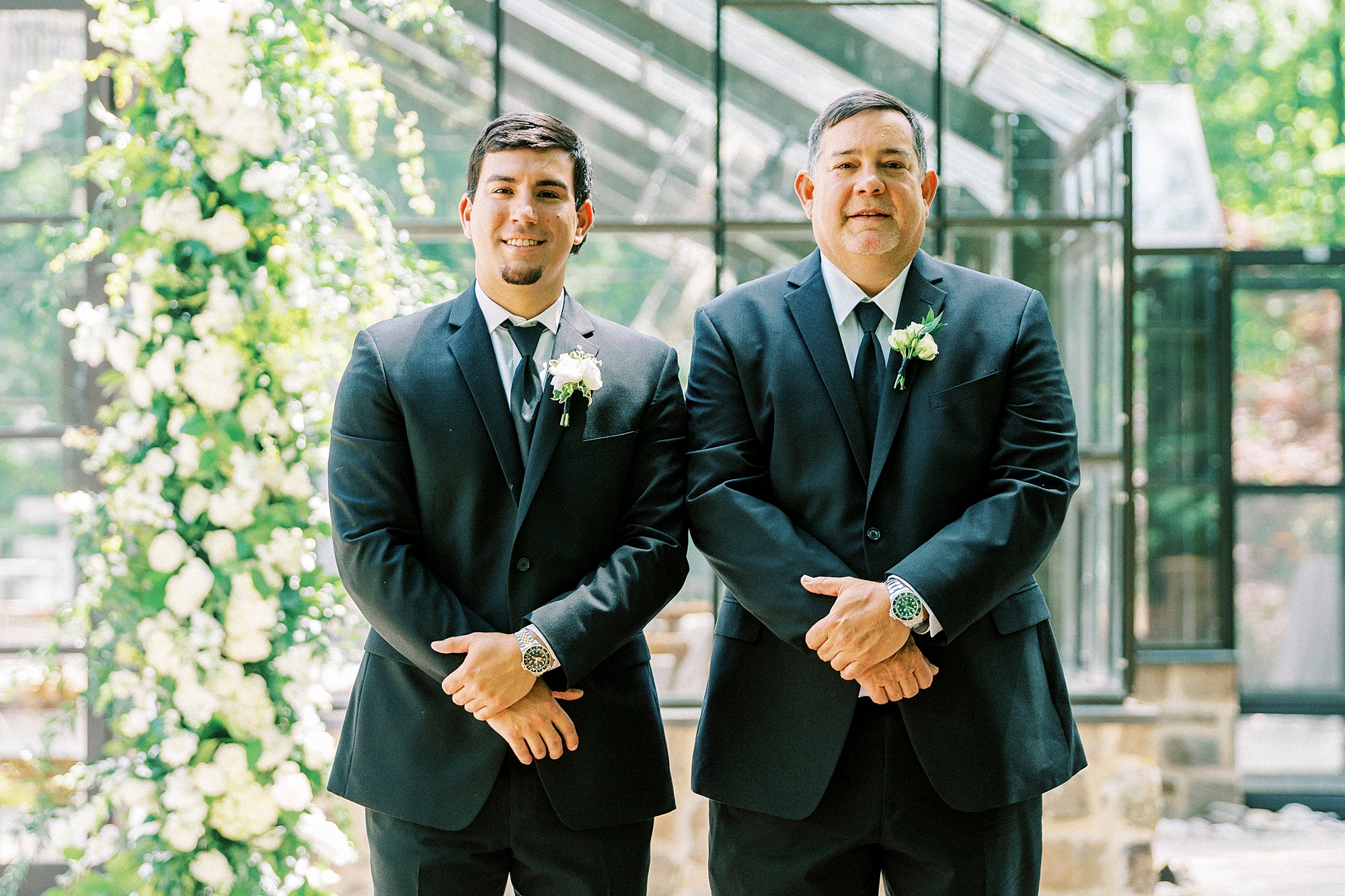 groom stands next to father in black suit by glass house at Morning Glory Farm