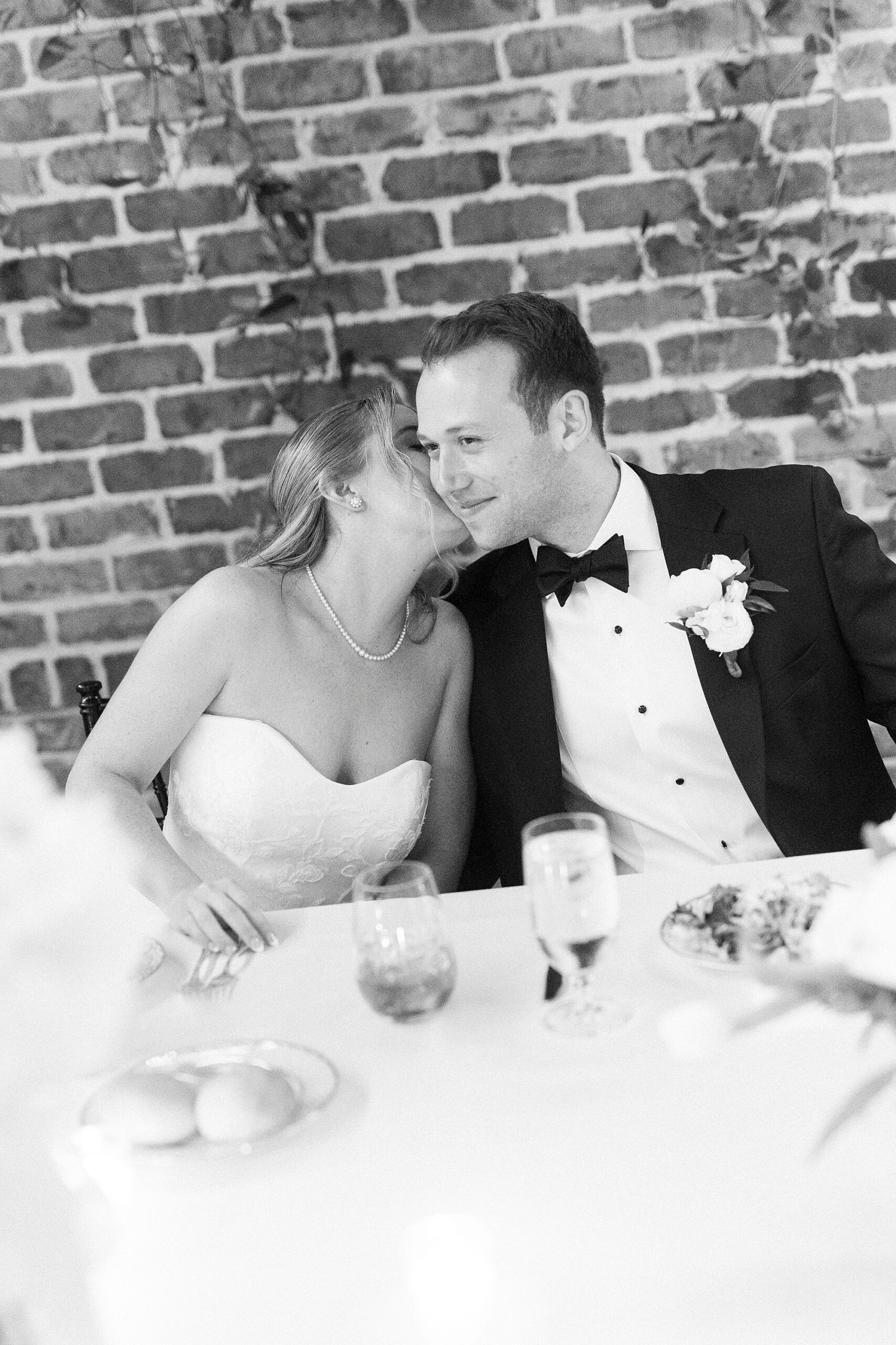 bride leans to kiss grooms's cheek sitting at reception