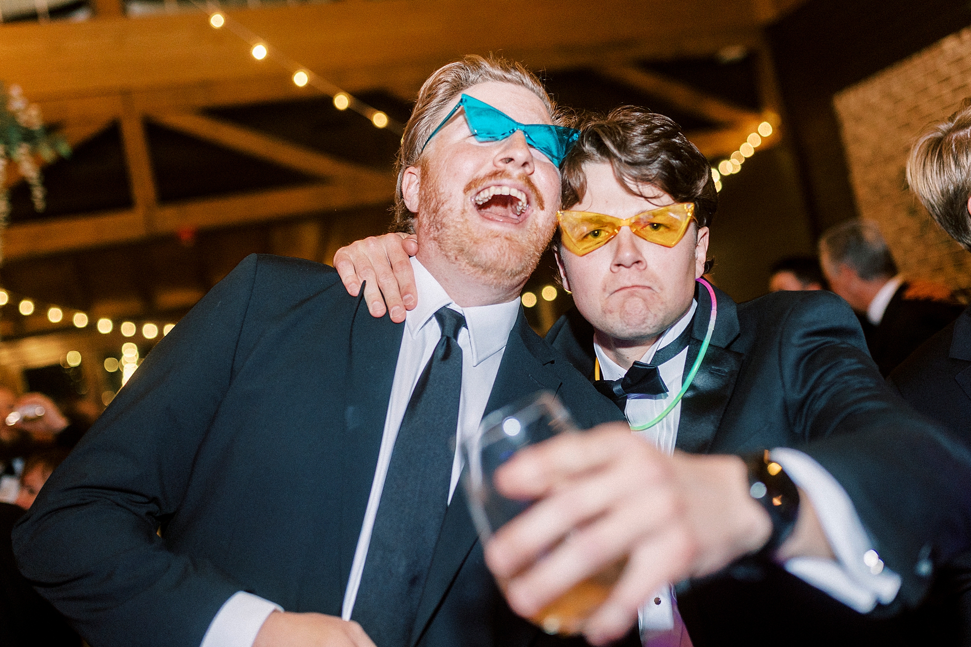 wedding guests in neon sunglasses laugh on dance floor during Wake Forest NC wedding reception in pavilion