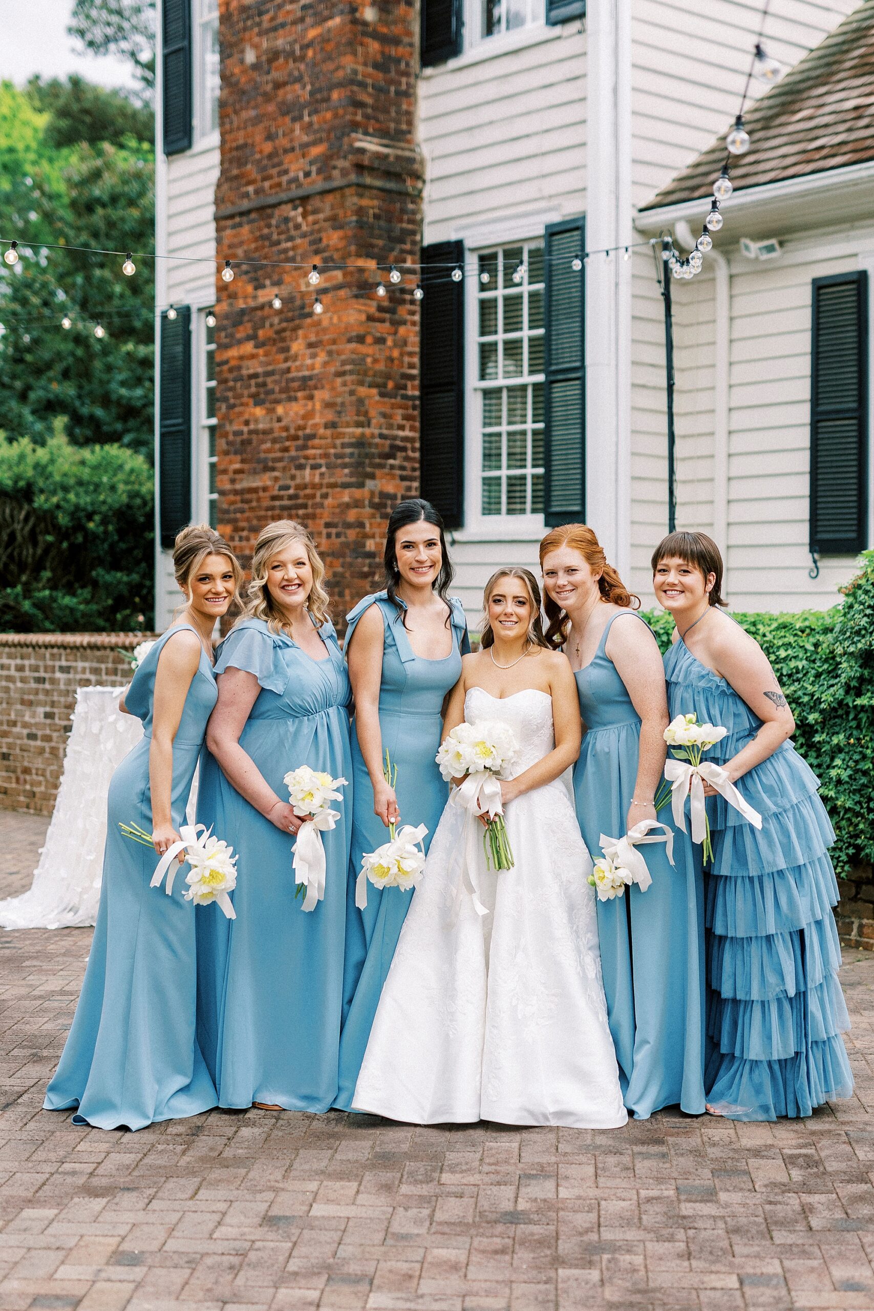 bridesmaids in mismatched light blue gowns hug around bride with white bouquets 