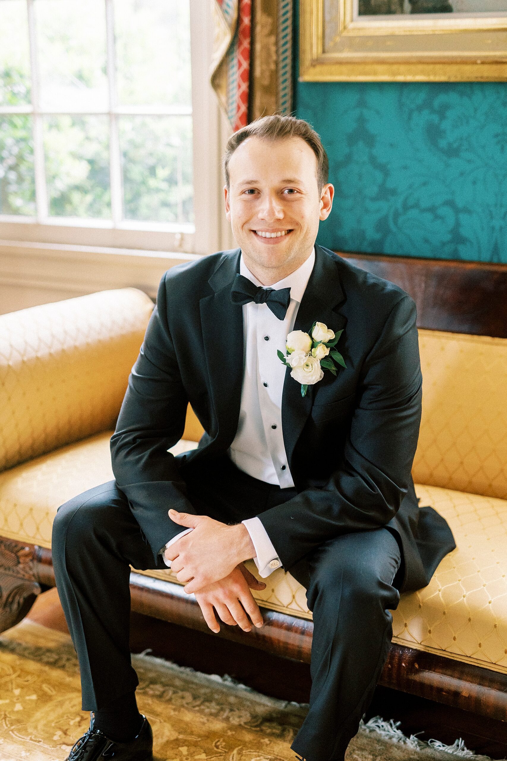 groom sits on leather sofa in black suit during wedding day prep
