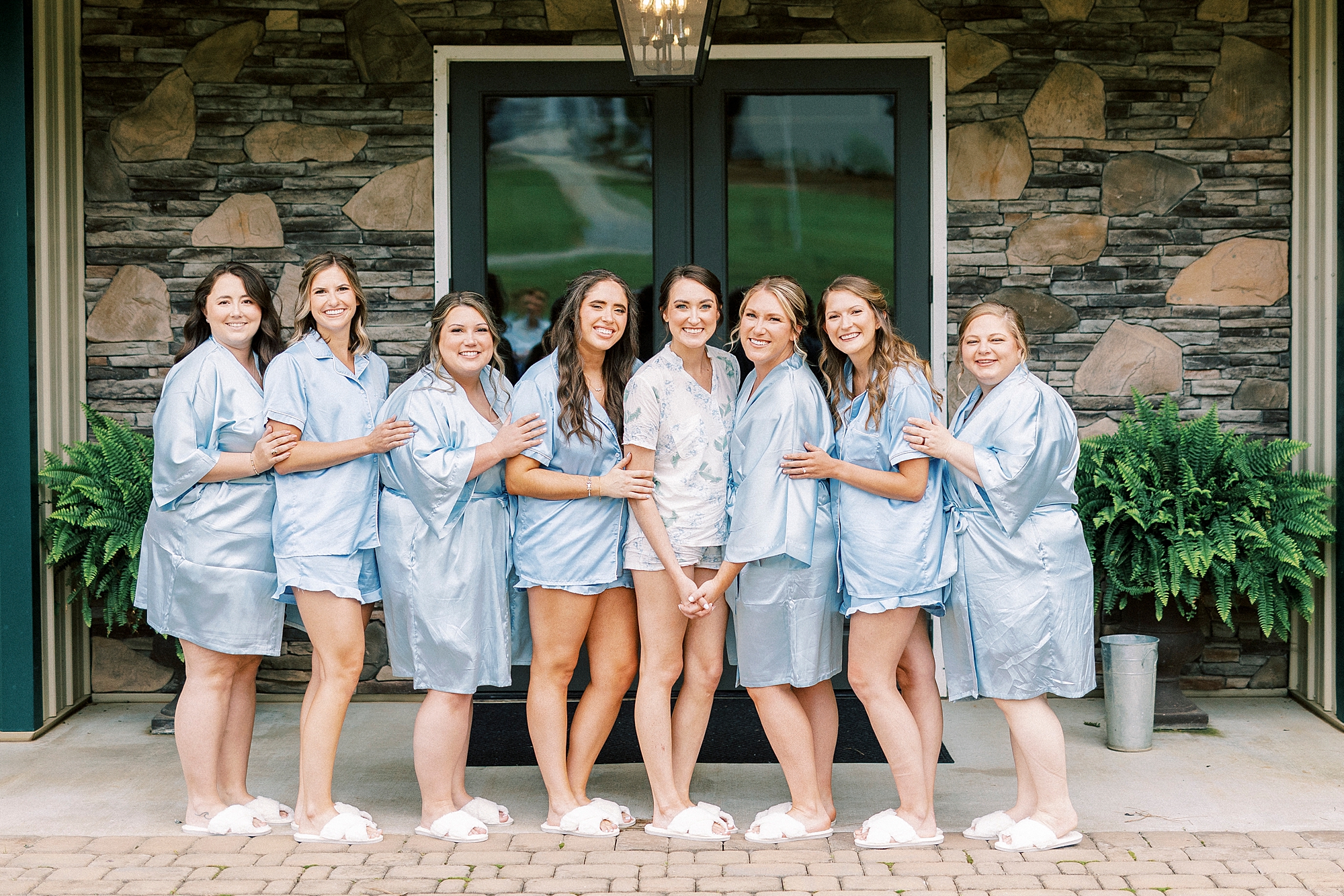 bride poses with bridesmaids in light blue robes outside stone house