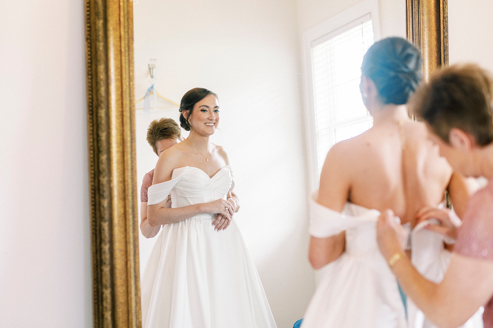 woman helps bride into wedding gown in reflection of mirror