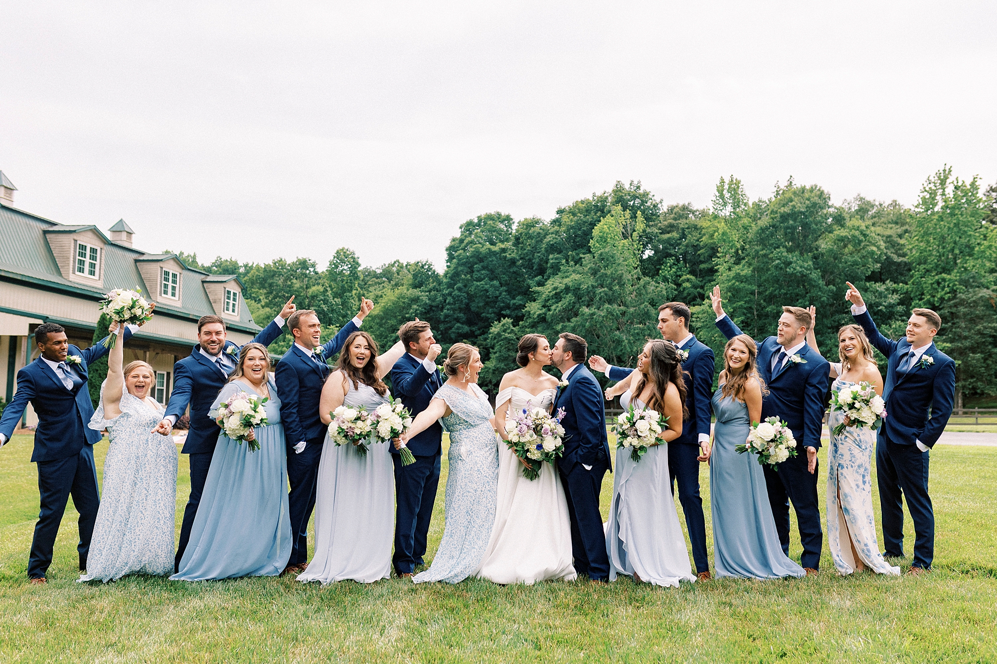 wedding party cheers while bride and groom kiss on lawn at Low Meadows Estate
