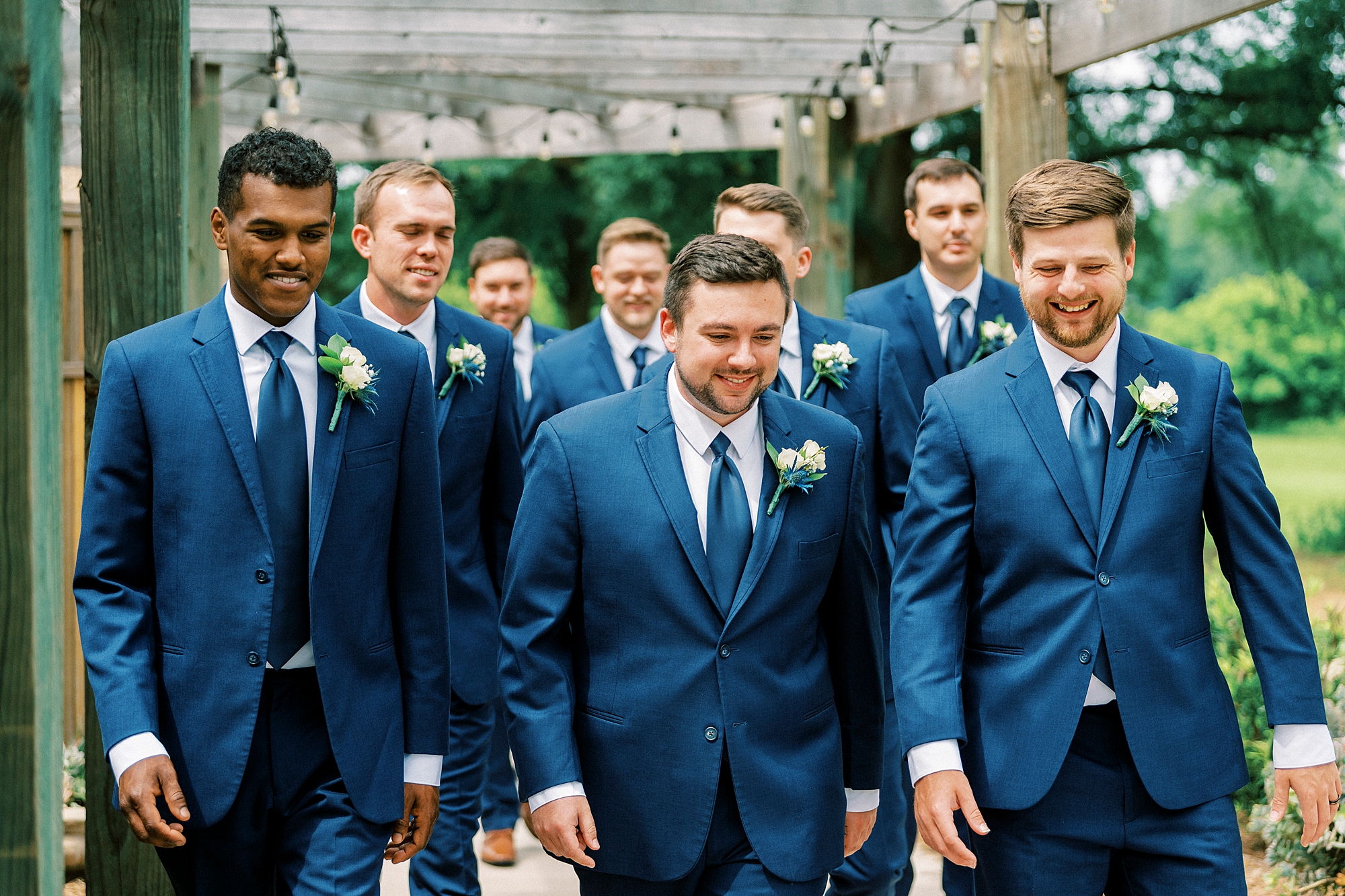 groom walks with groomsmen in blue suits through gardens 