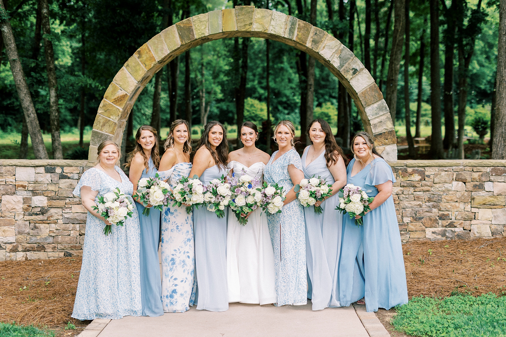 bride stands with bridesmaids in mismatched light blue gowns in gardens of Low Meadows Estate