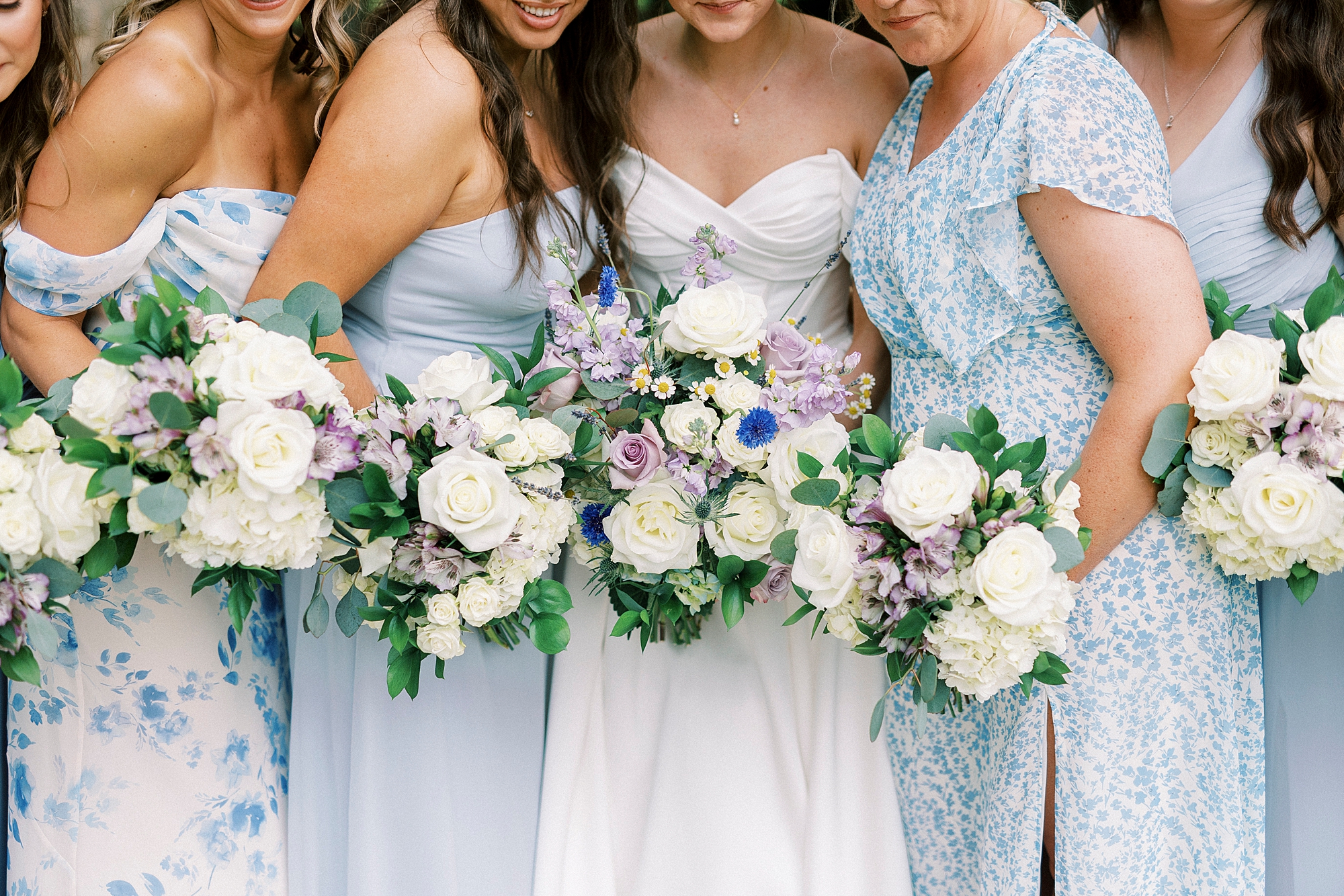bride and bridesmaids stand together holding light purple and white bouquets 
