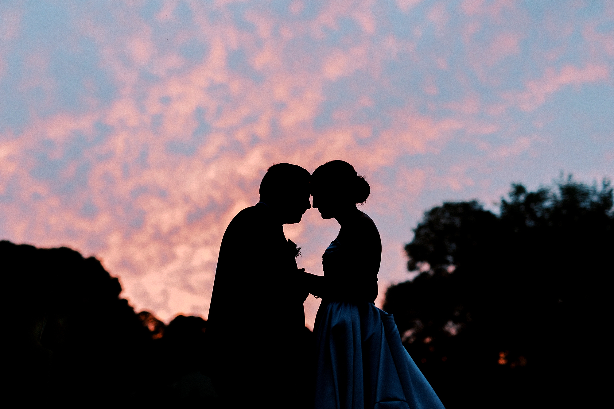 bride and groom lean heads together silhouetted in front of pink and blue sky