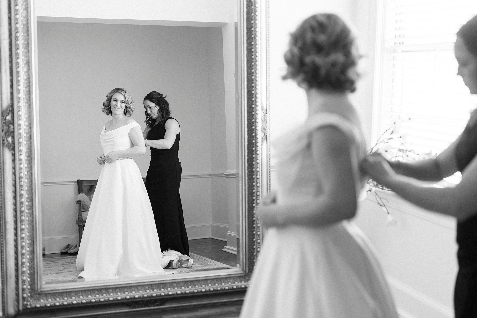 bridesmaid helps bride into wedding gown in reflection of mirror in bridal suite 