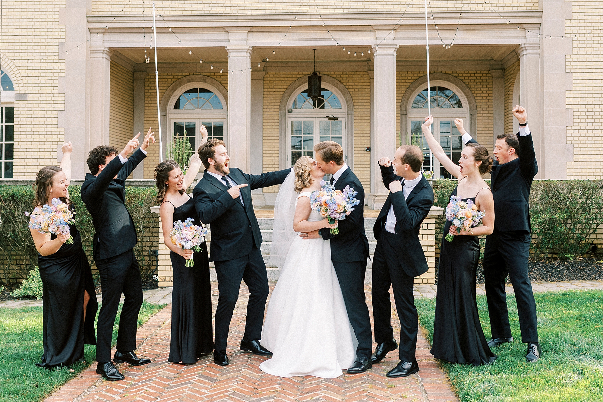bride and groom kiss with wedding party in black cheering around them