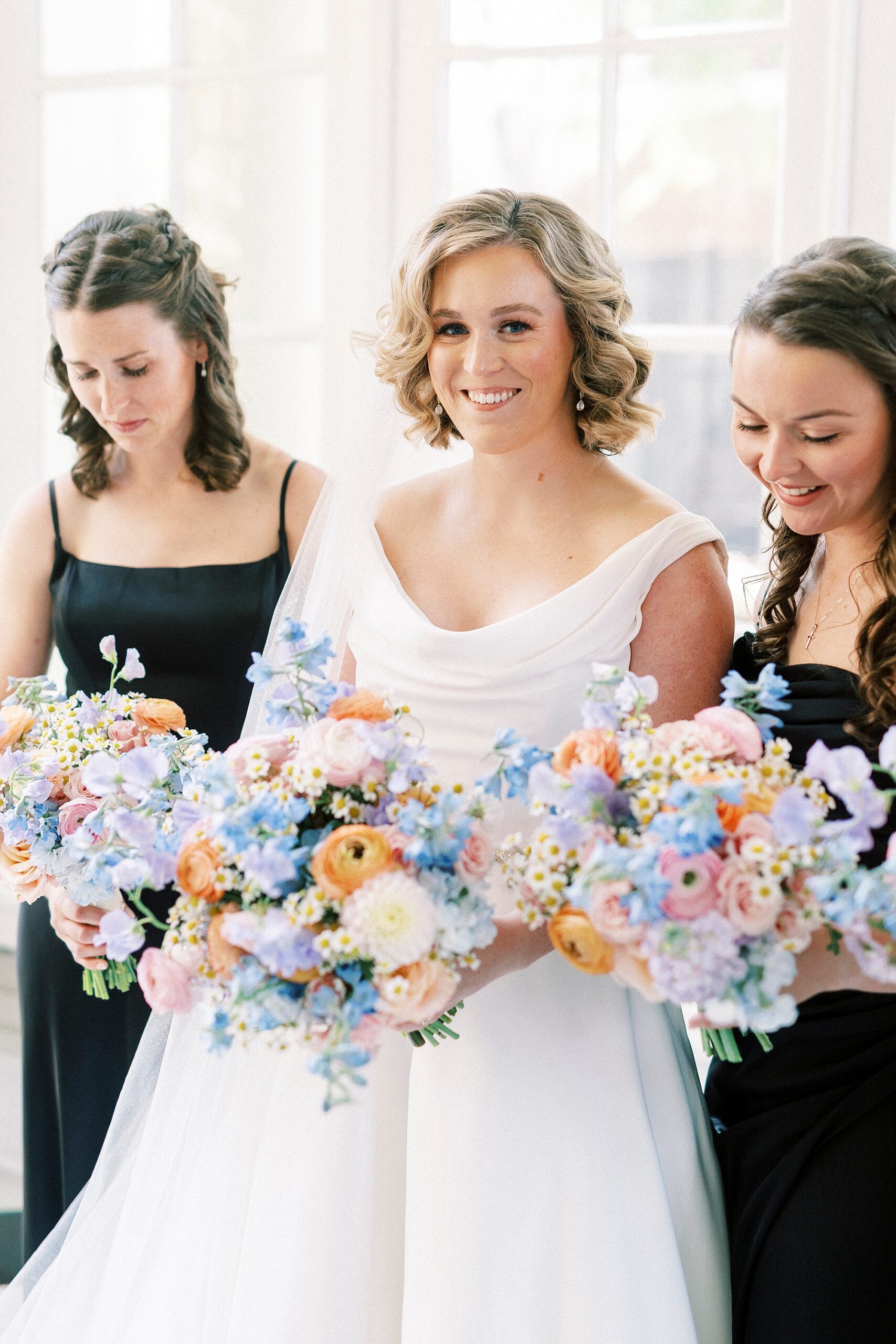 bridesmaids in black gowns pose with bride holding bouquets of pastel flowers 