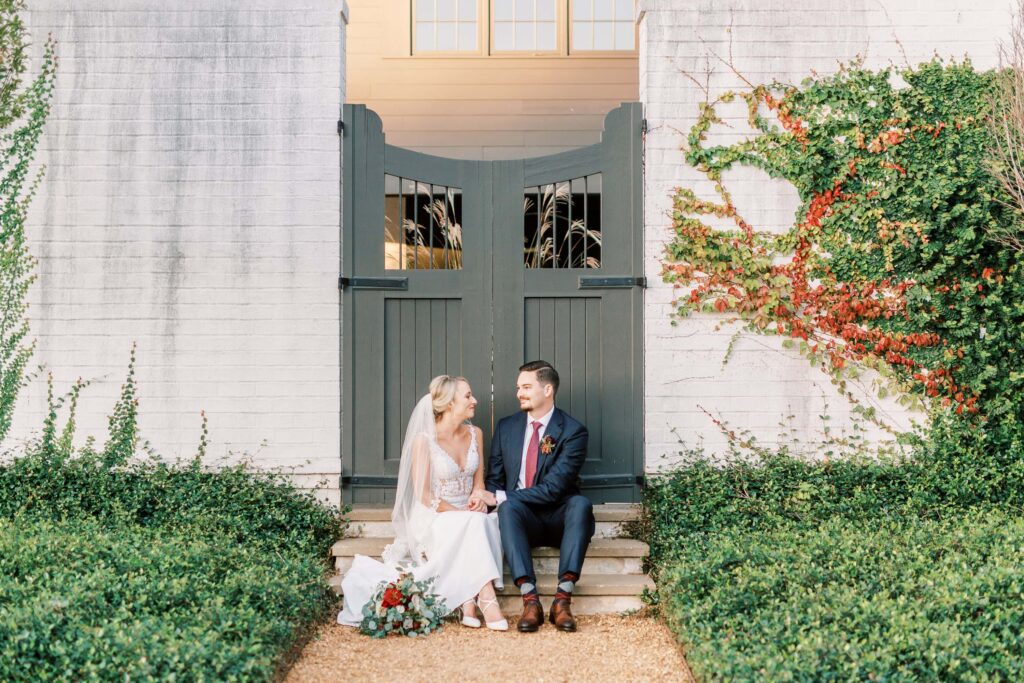 bride and groom sit on steps in front of gate and greenery at the Andrews farm in midland North Carolina
