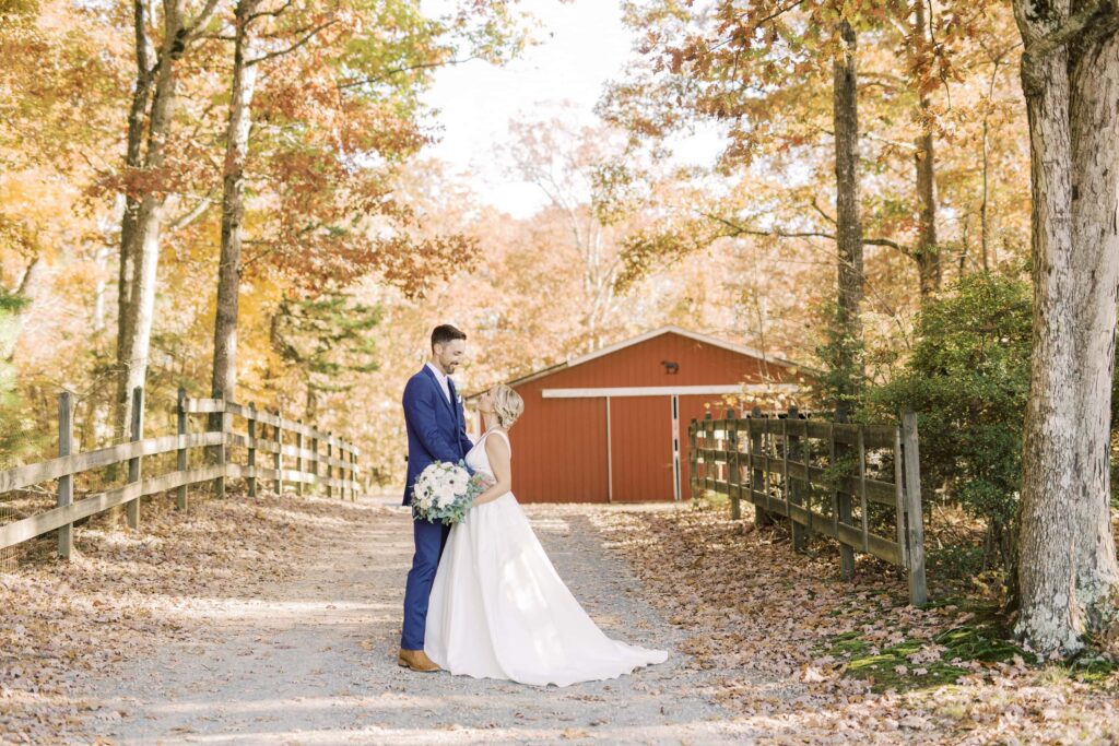 bride and groom embrace and smile on path with red barn and fall leaves at the farm at brusharbor