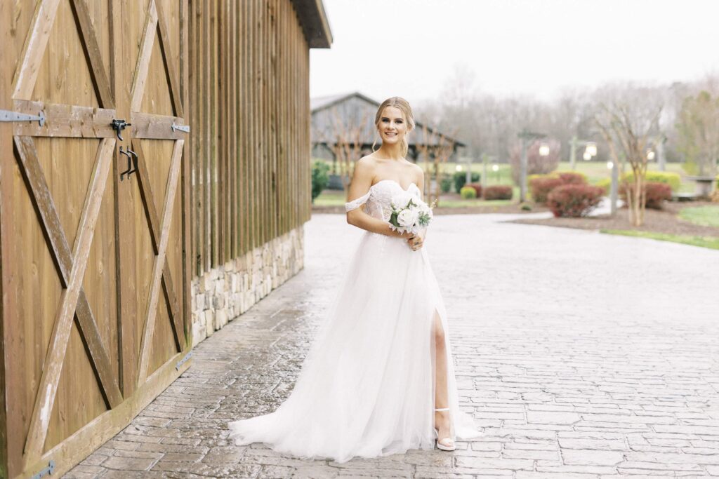 bride smiles and holds simple bouquet in front of brown barn at the farmstead wedding venue
