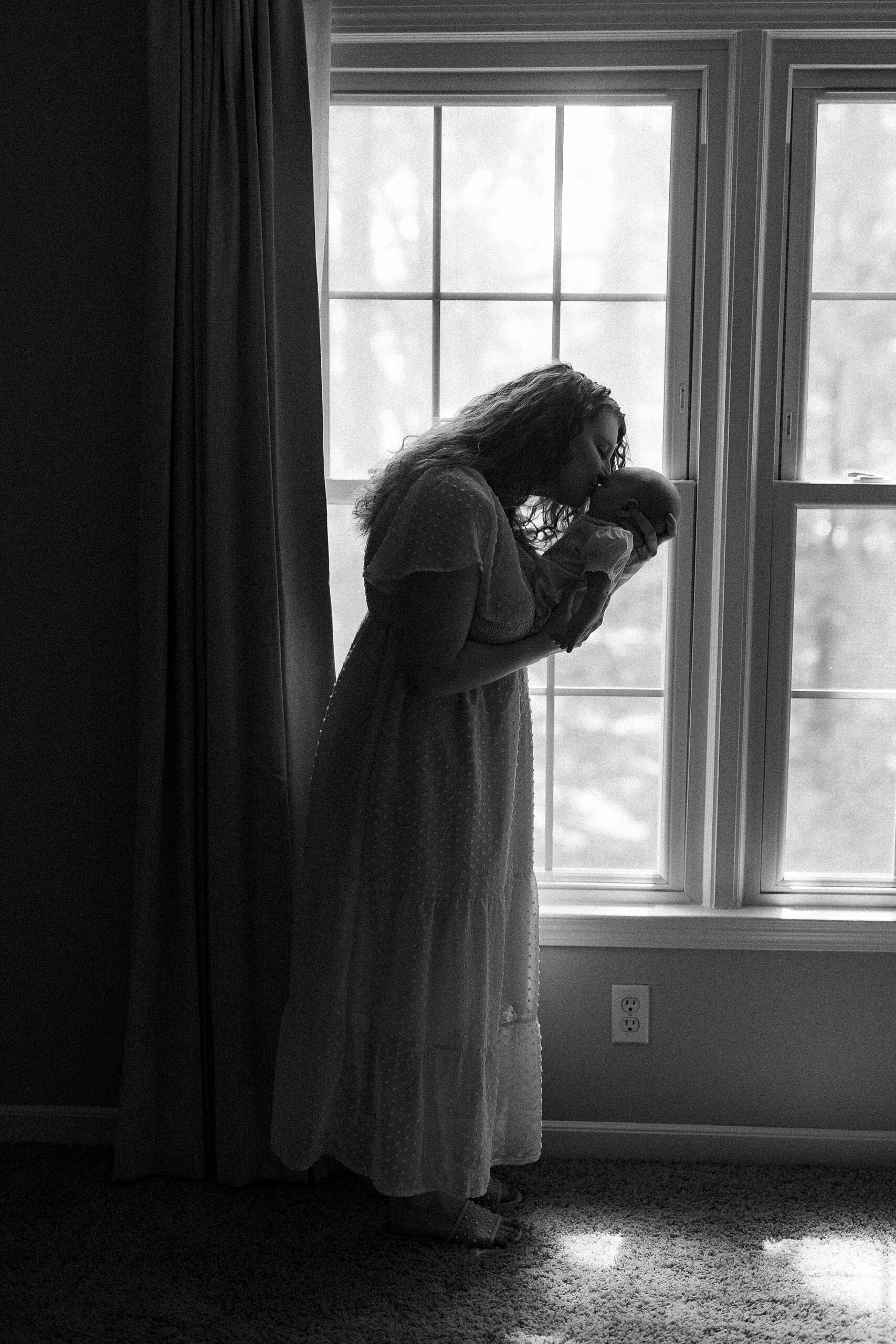 black and white photo of mom leaning down to kiss baby in window 