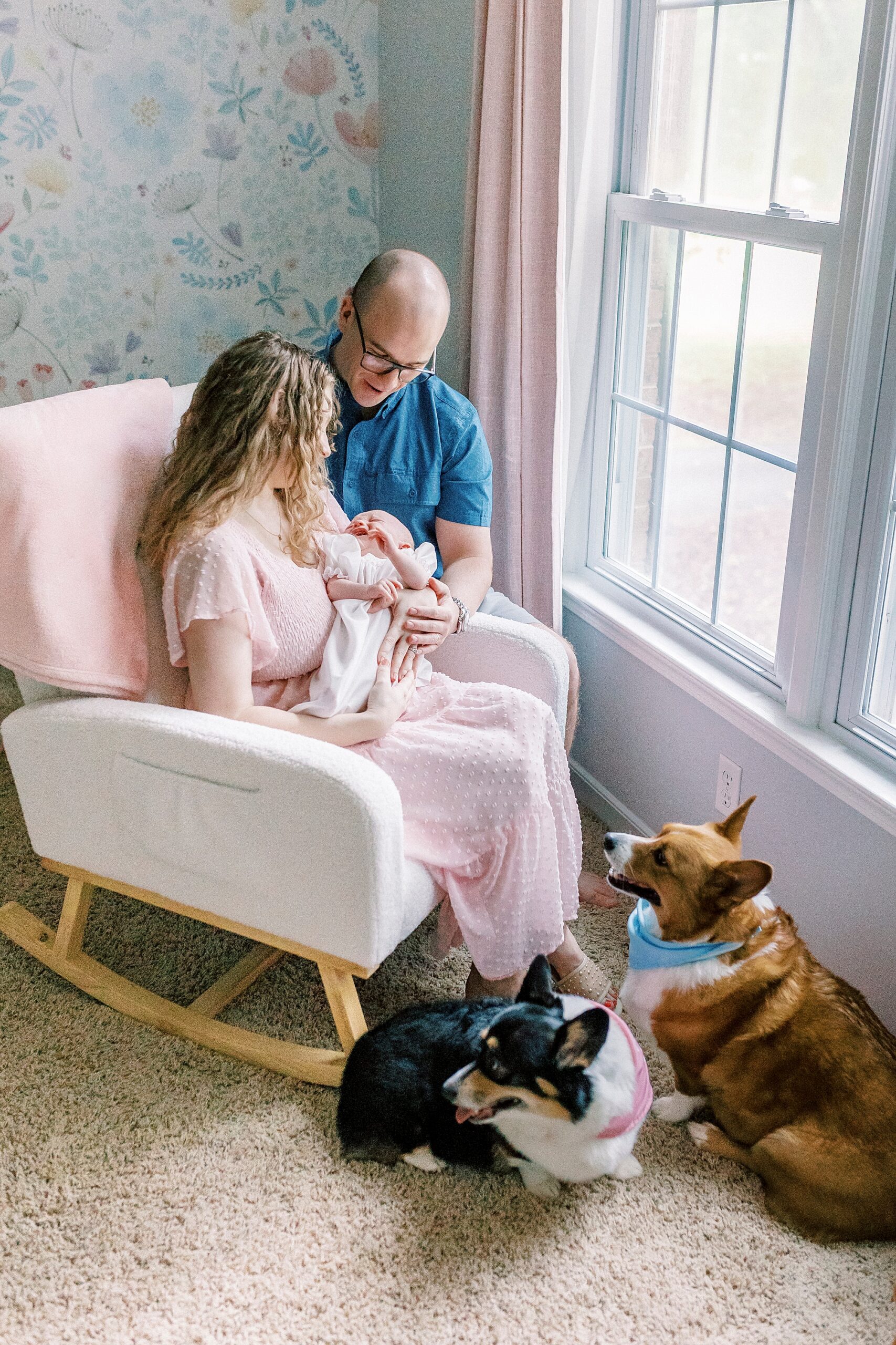 mother holds baby girl in rocking chair by window while dad looks over their shoulder 