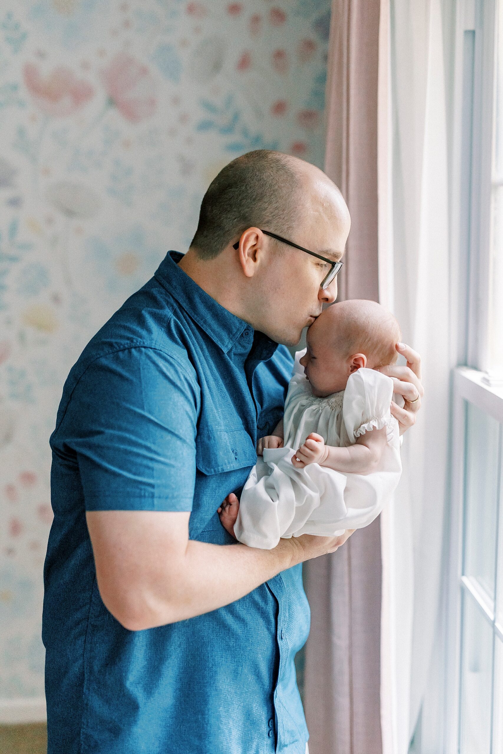 dad holds newborn daughter by window kissing her forehead 