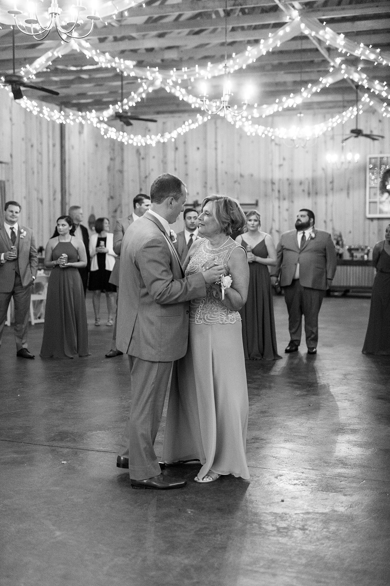 groom dances with mother during wedding reception in barn with twinkle lights hanging