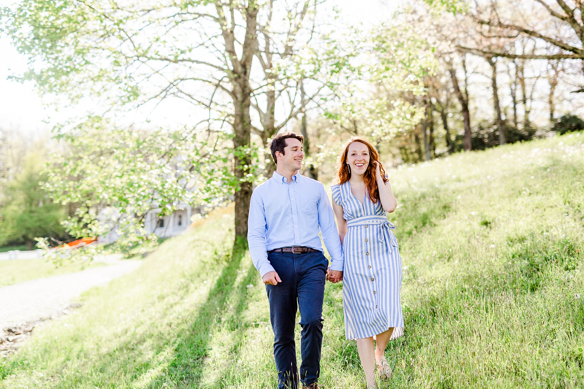 couple walks through Moses Cone Manor grounds during spring engagement photos in blue dress and blue shirt