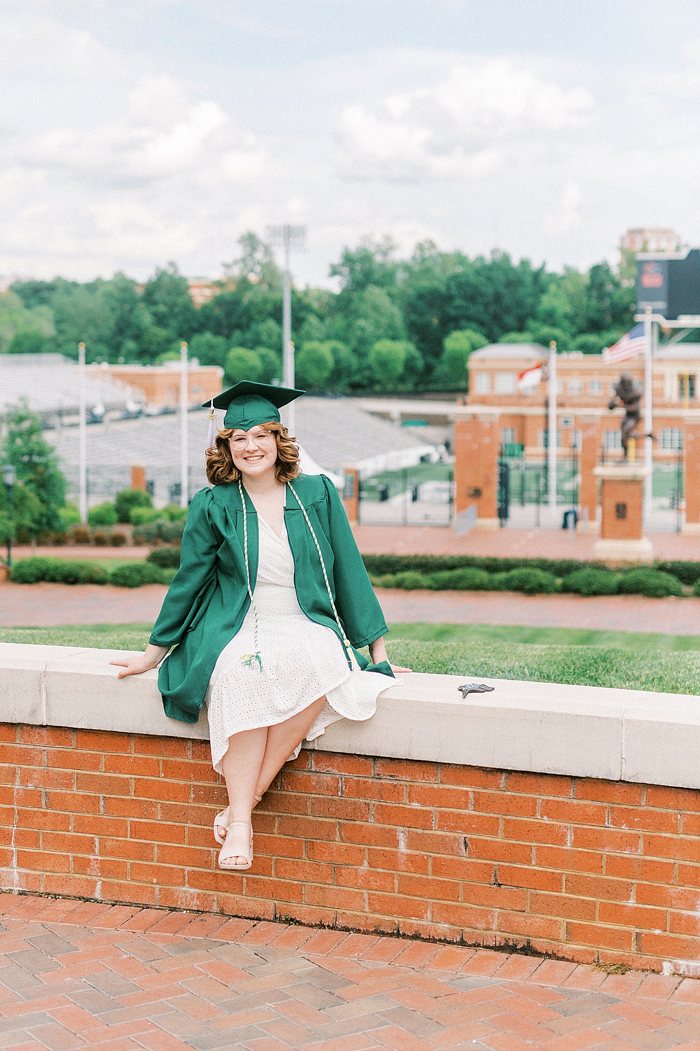 college graduate sits on brick wall by football field at UNCC