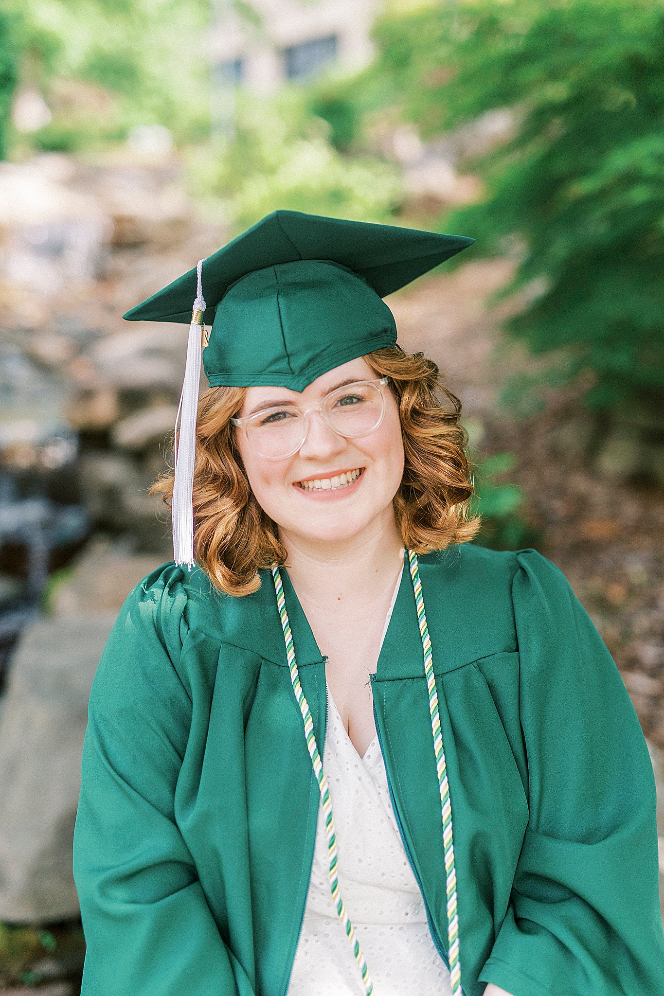senior poses in green cap and gown by water in Charlotte NC