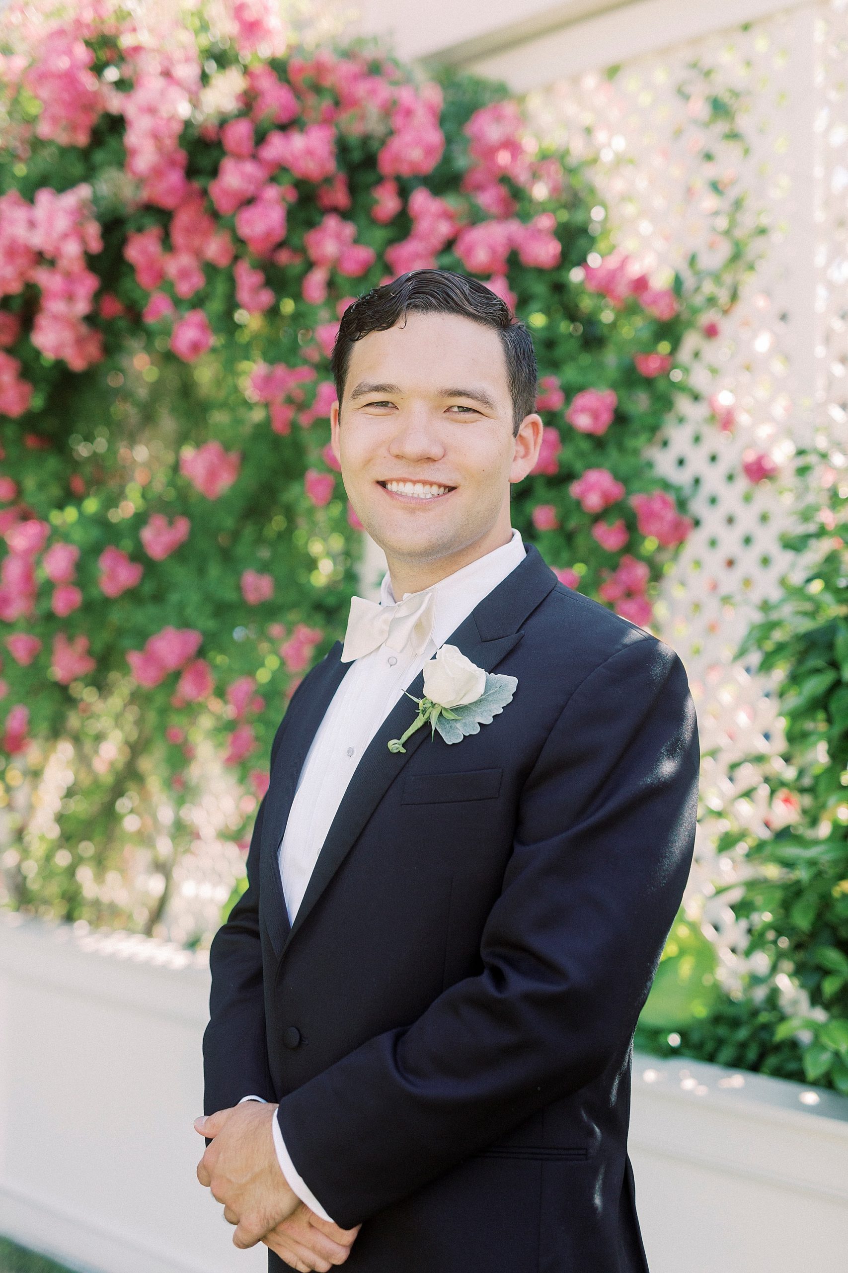 groom in classic tux with white boutonniere poses by pink flowers