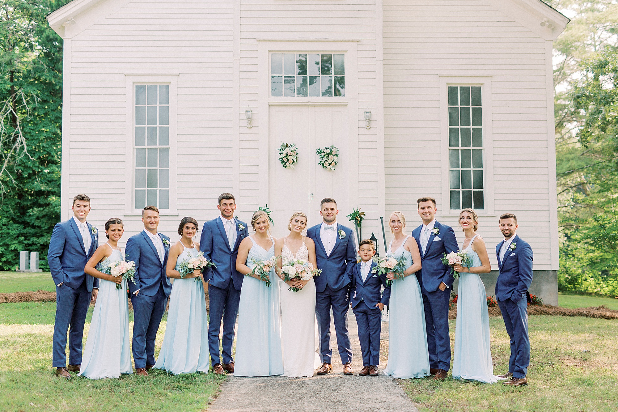 newlyweds pose with wedding party outside little white church