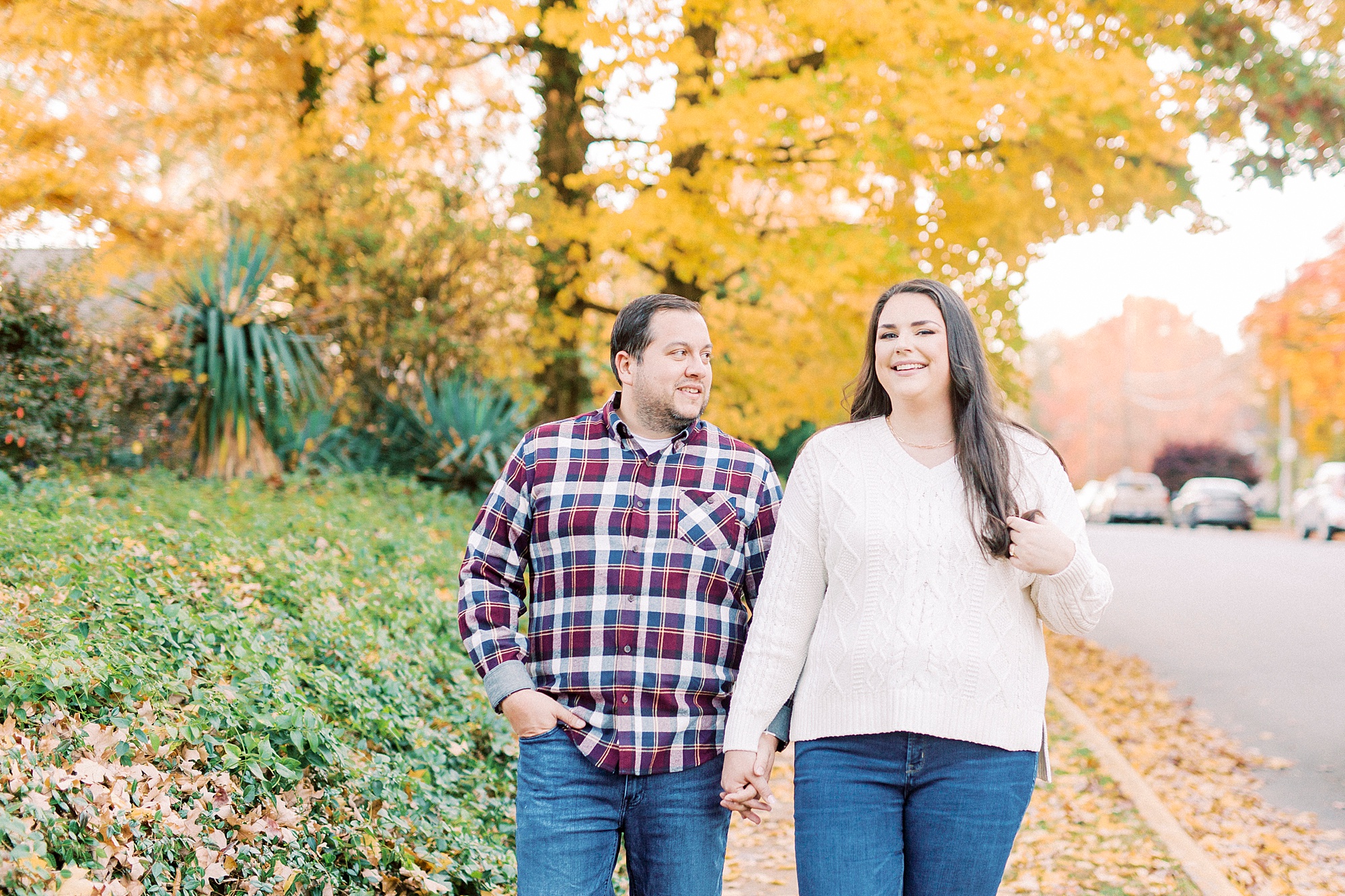 engaged couple walks holding hands on sidewalk with yellow leaves behind them