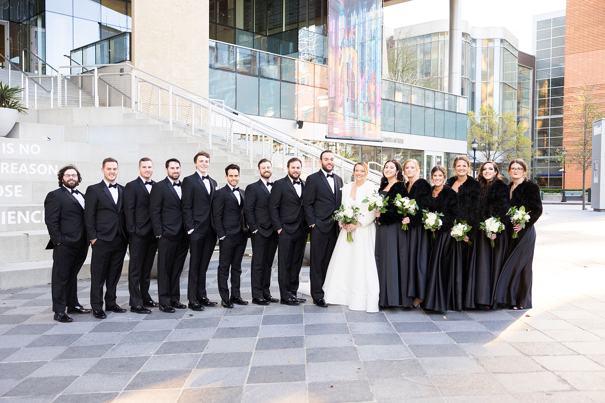 newlyweds pose outside Mint Museum Uptown with bridal party in all black