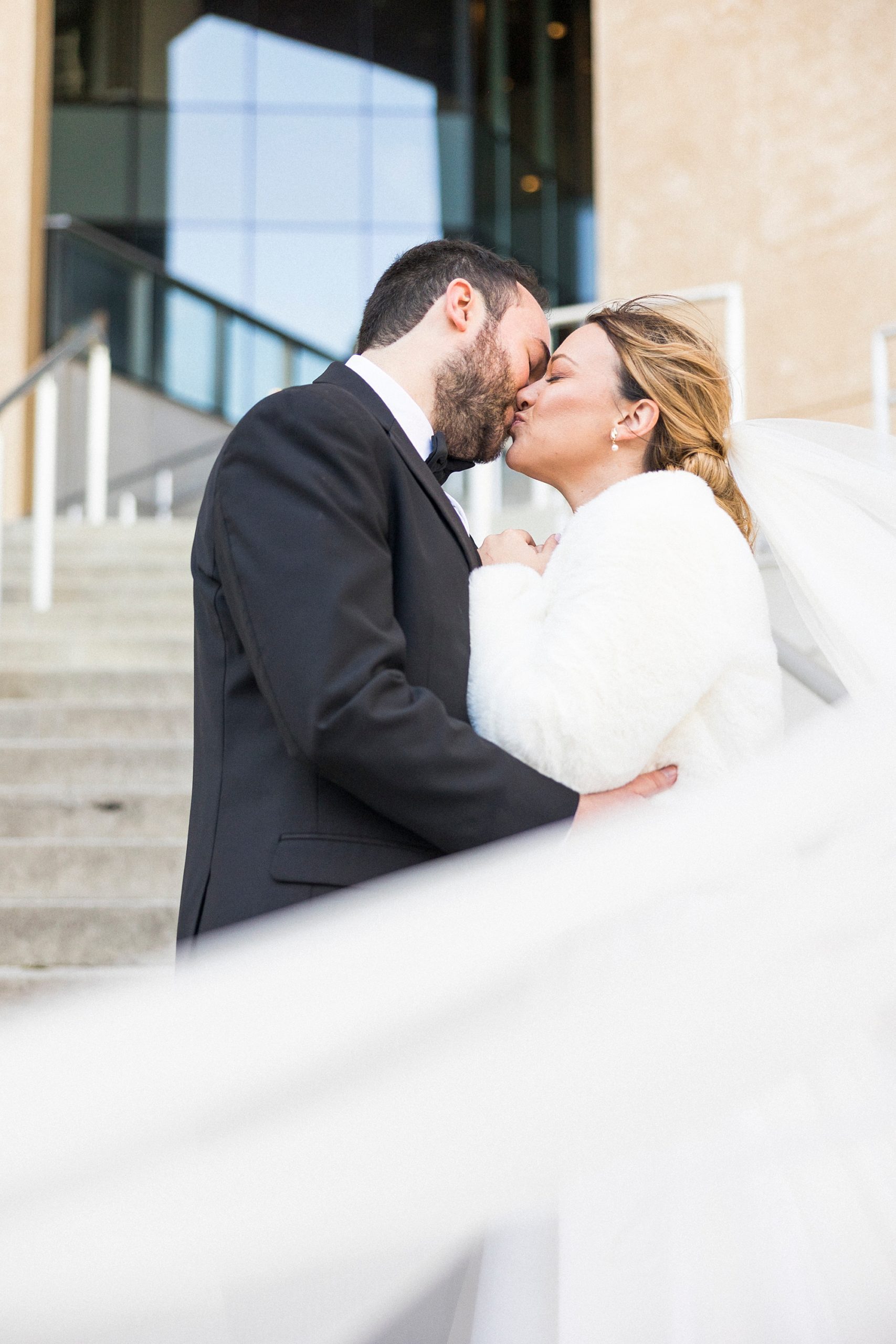 newlyweds kiss on steps of Mint Museum Uptown with bride's veil floating