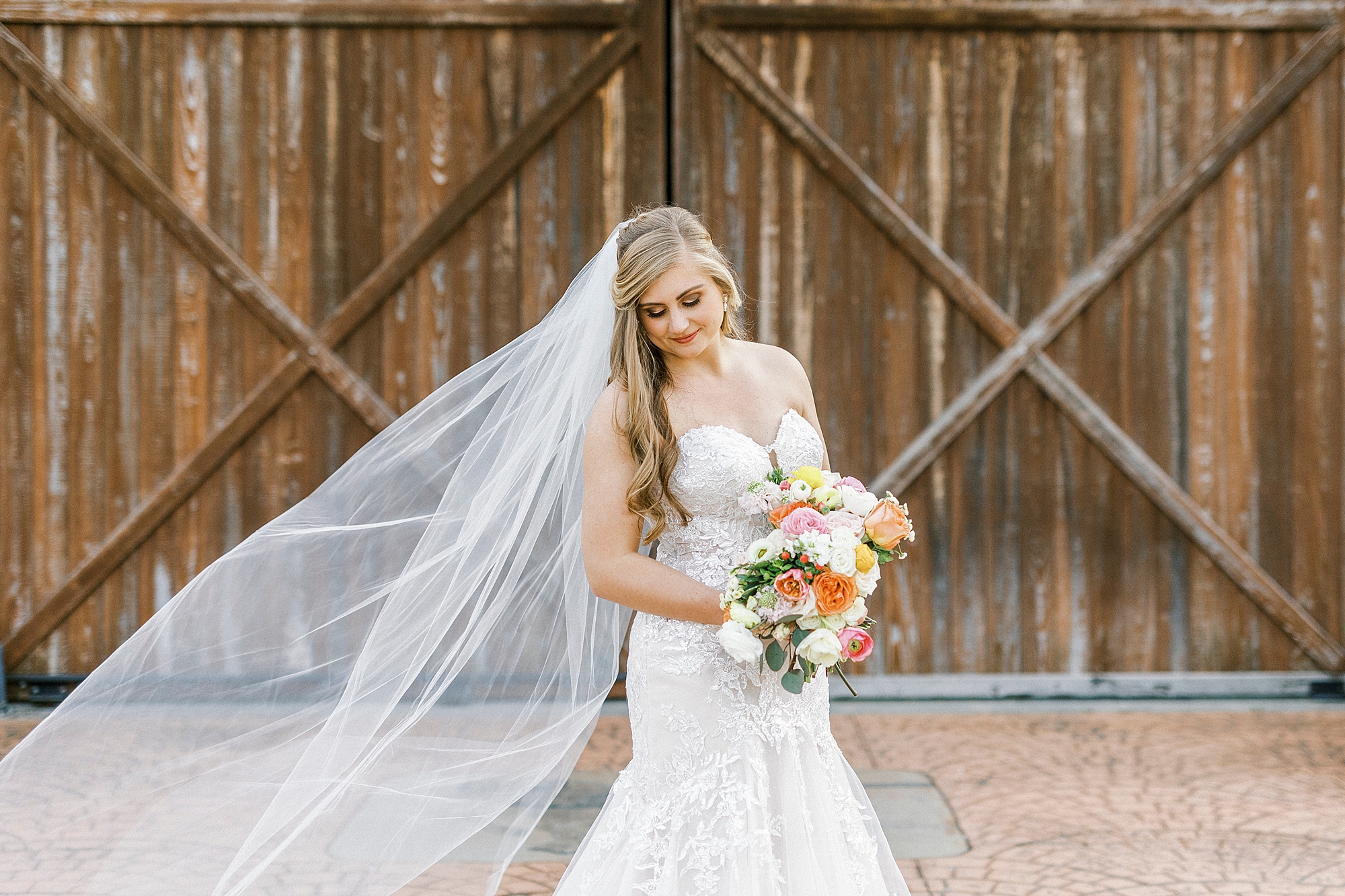 bride holds bouquet of colorful flowers standing by wooden doors of The Stable at Riverview