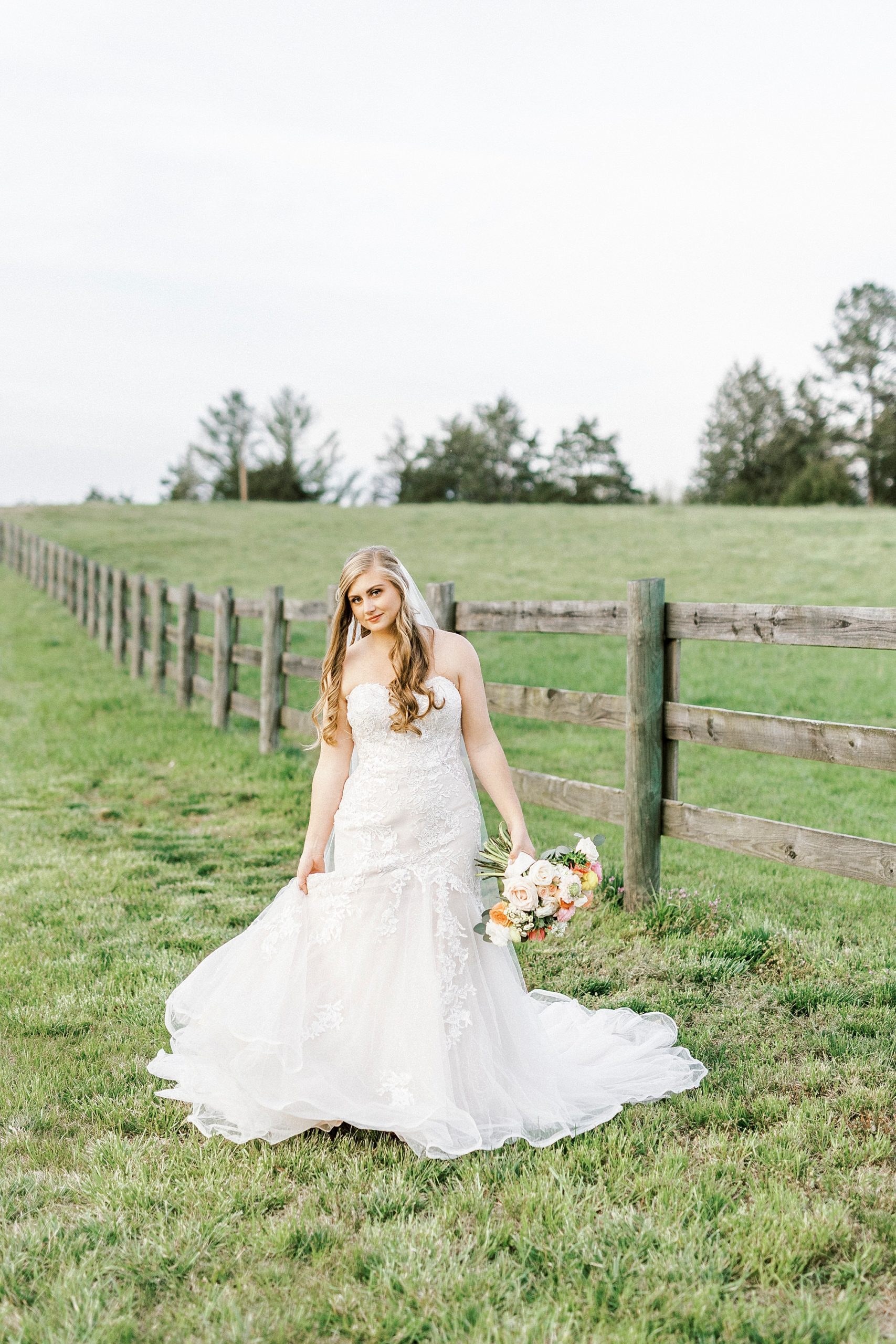 bride walks in grass along wooden fence at The Stable at Riverview