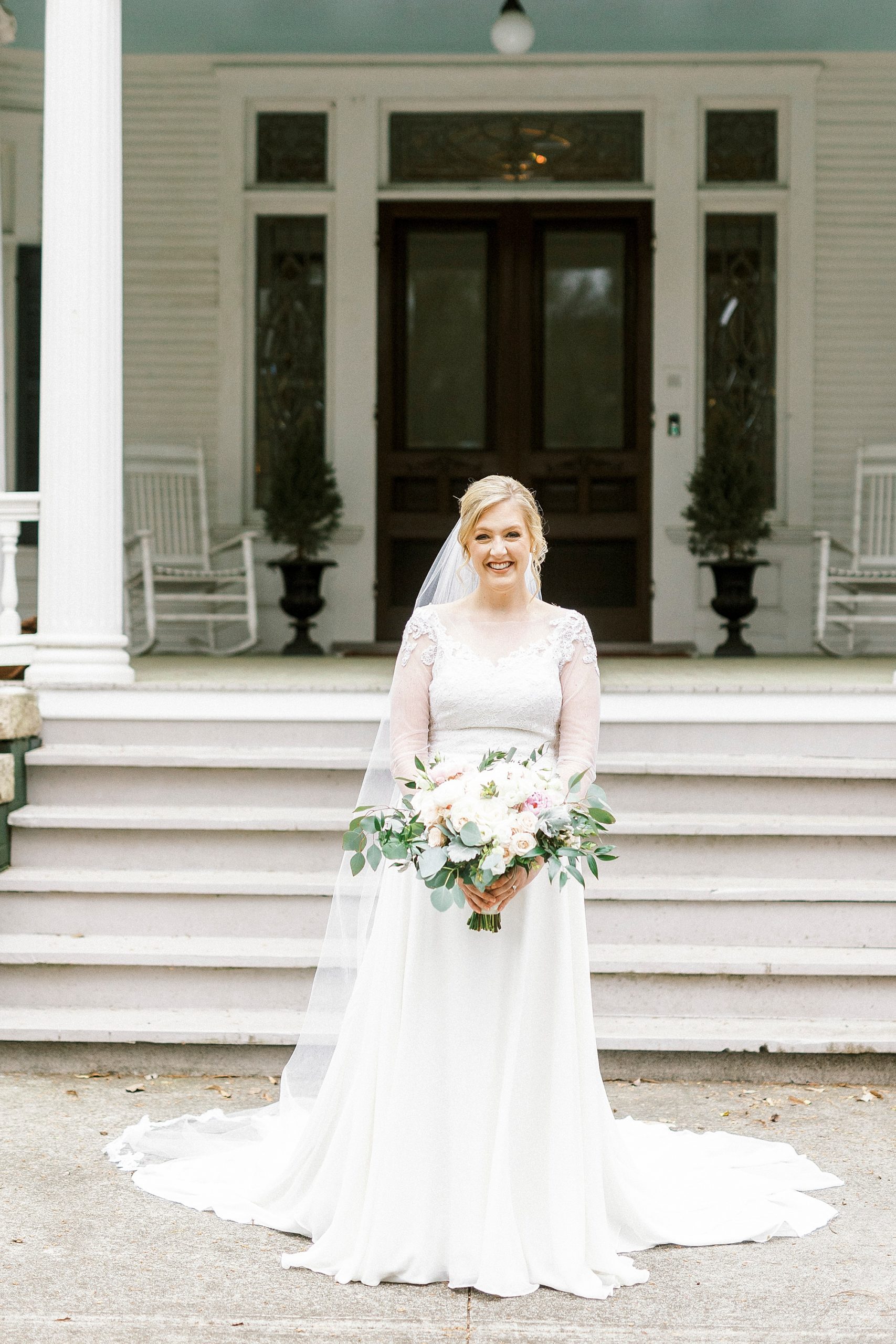 bride stands outside historic home with bouquet of white and pink flowers