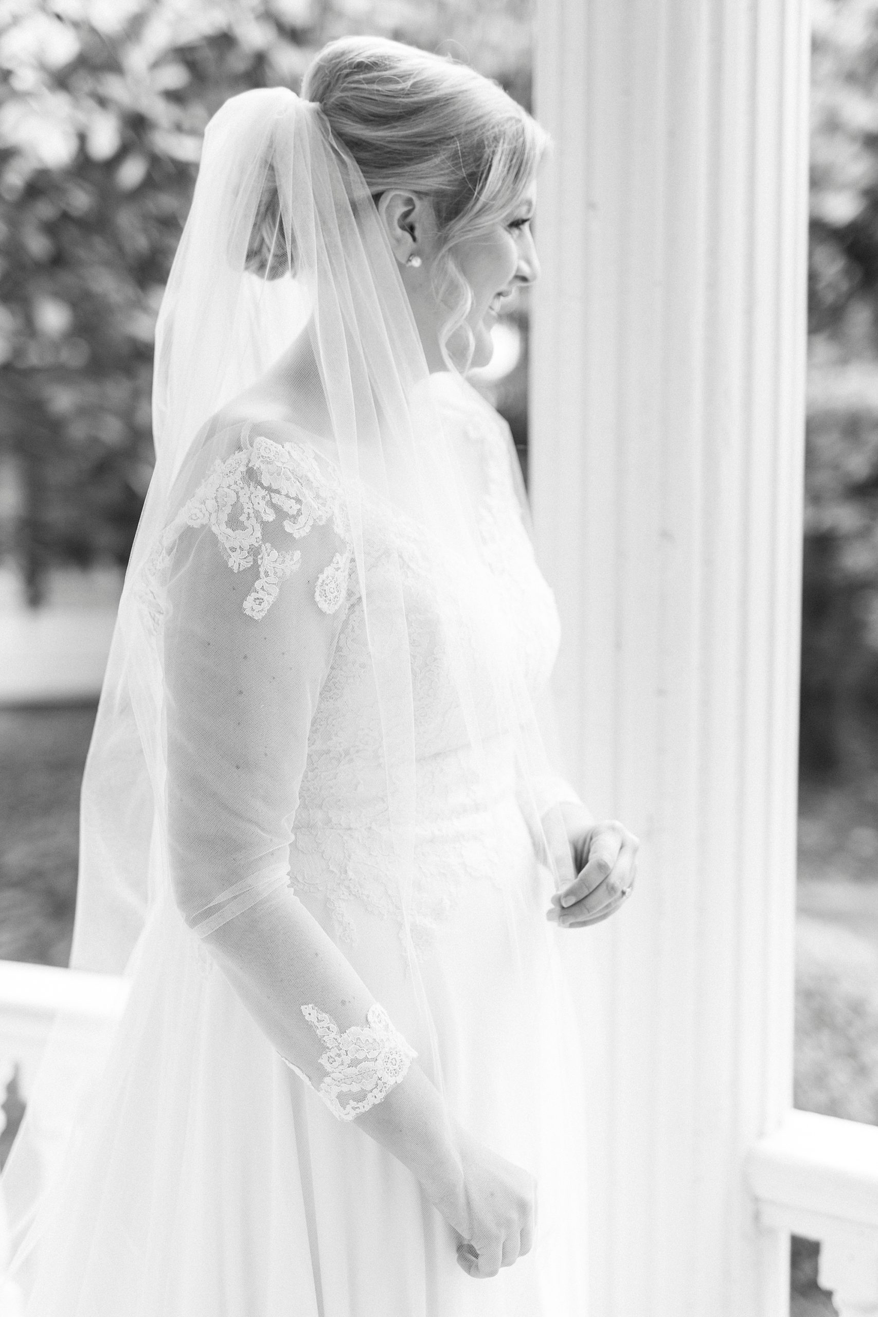 bride stands on front porch looking at yard with veil around shoulders 