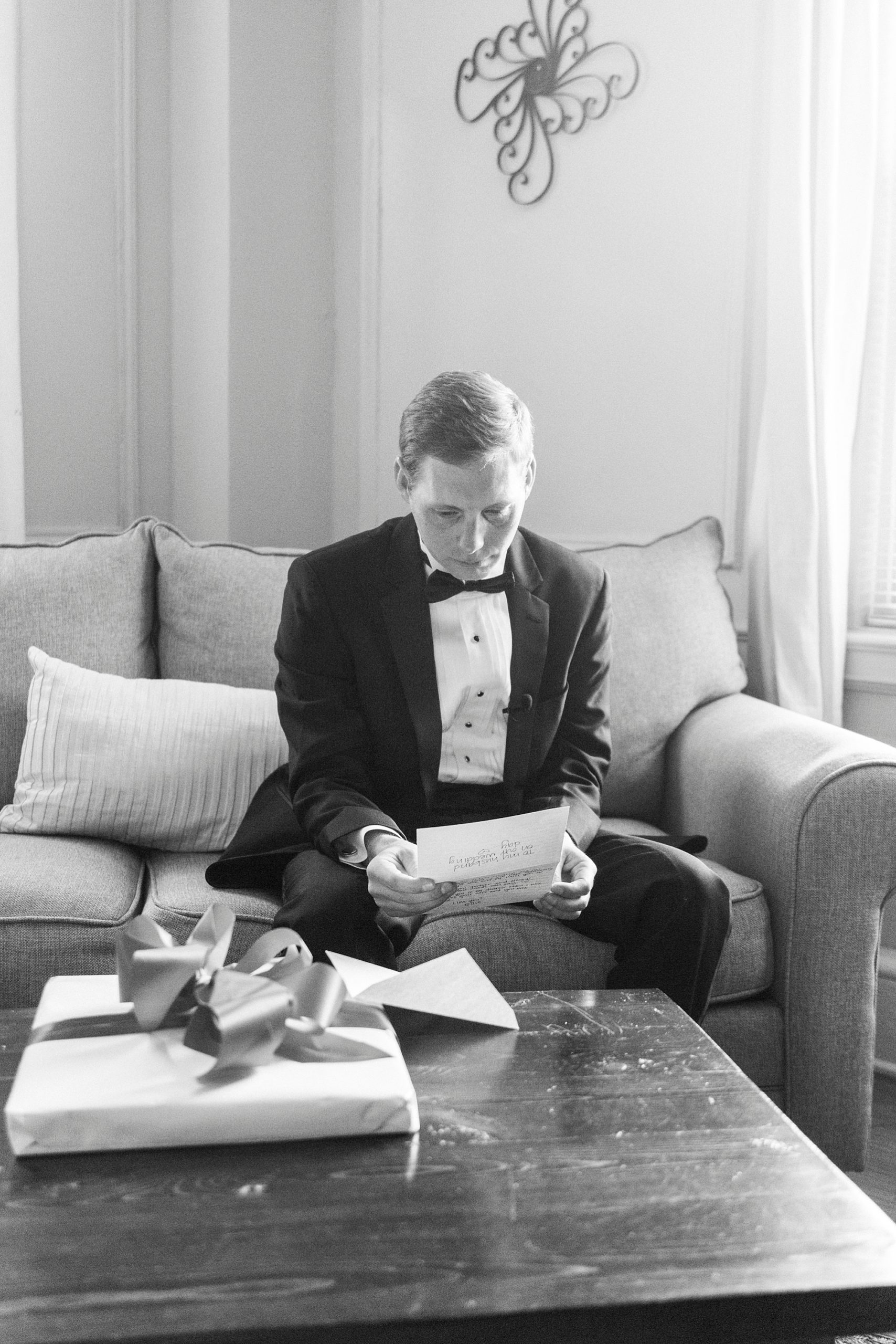 groom sits on couch opening gift on wedding day