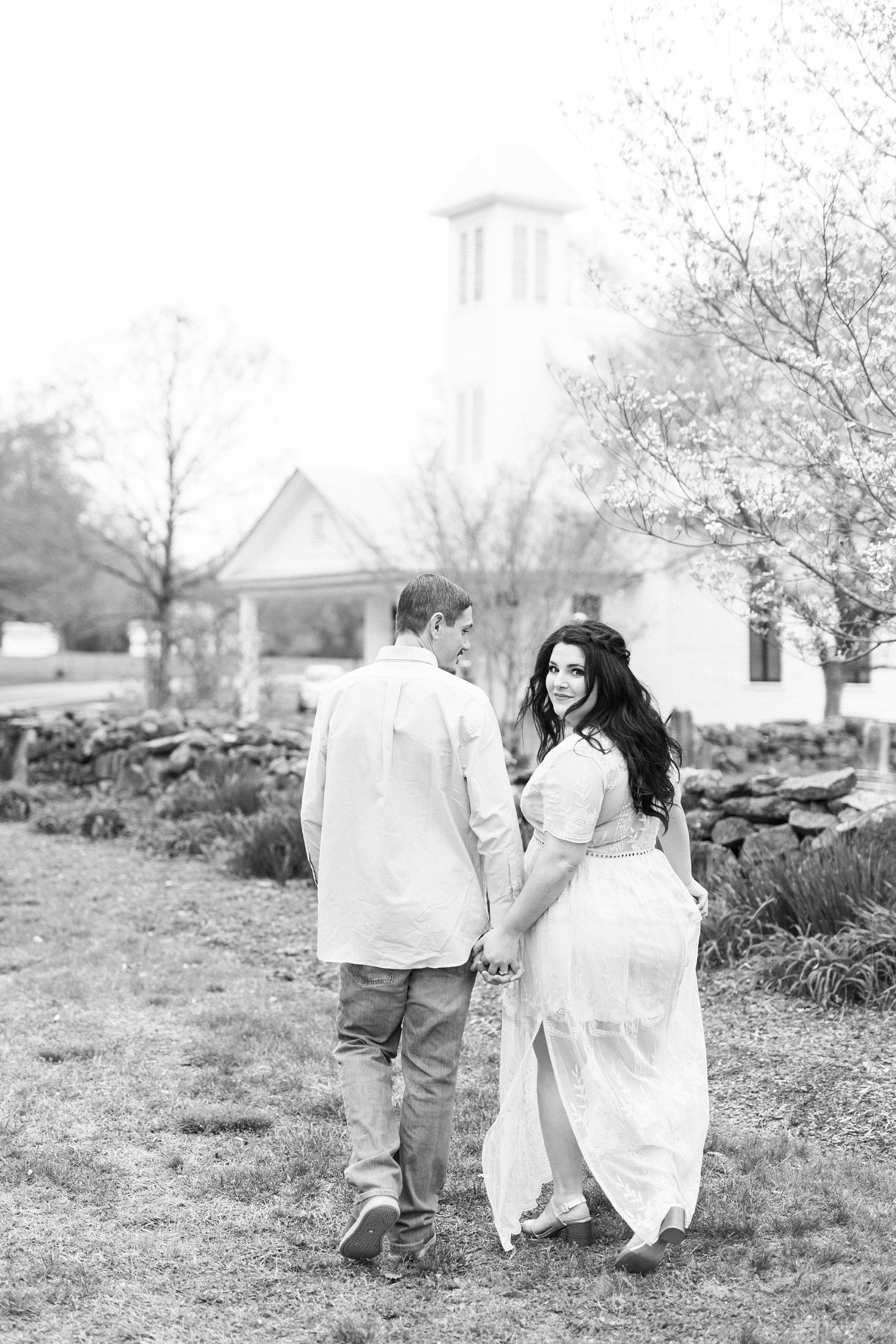bride and groom hold hands walking by stone wall during white chapel engagement session
