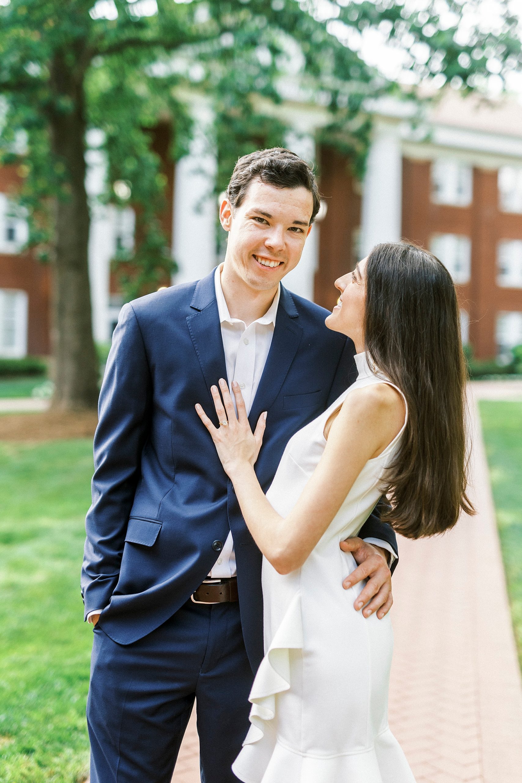 bride smiles up at groom while he looks at camera on pathway at Queens University