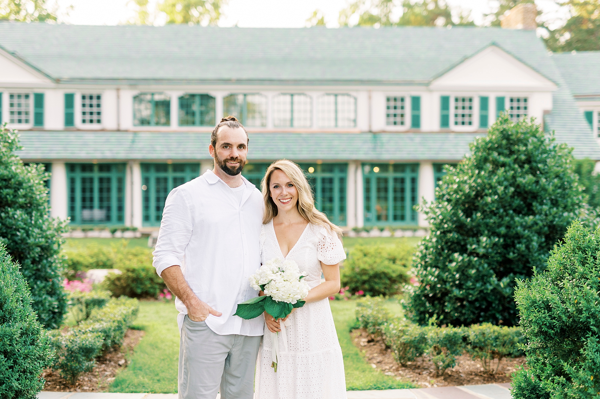 engaged couple stands outside Reynolda Gardens holding bouquet of white flowers 