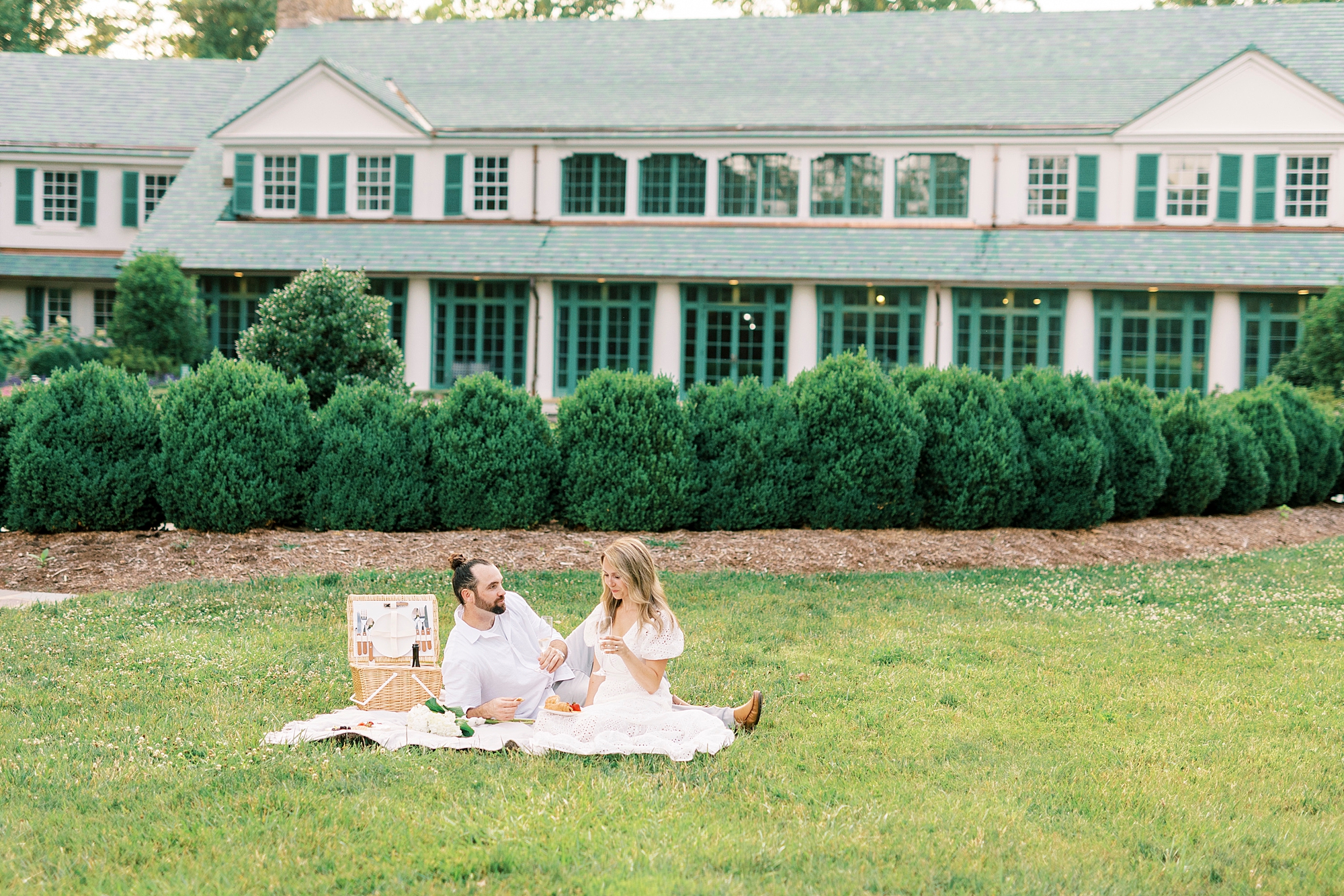 engaged couple sits on lawn of Reynolda Gardens with picnic 