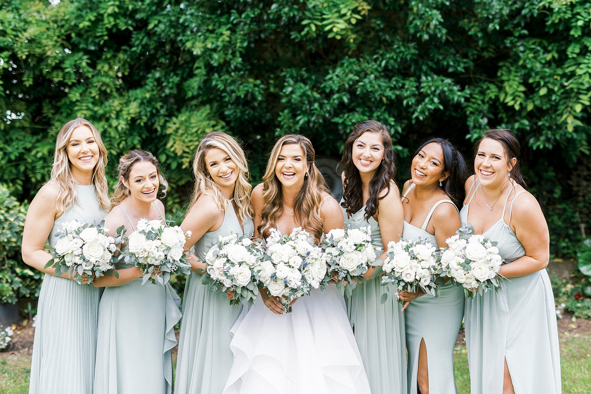 bride stands with bridesmaids in light green gowns holding all-white bouquets 