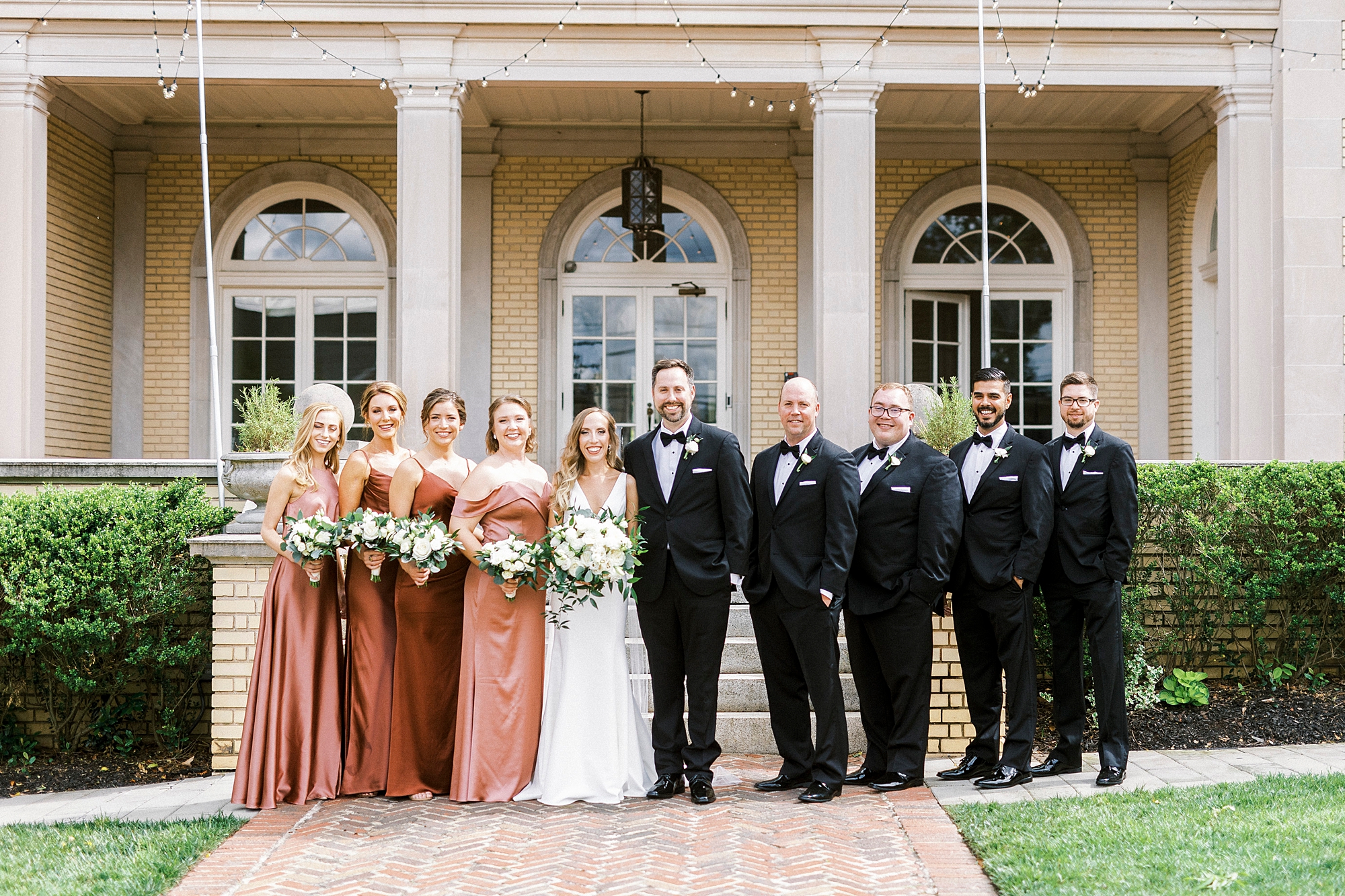 bride and groom stand with wedding party in copper gowns and black tuxes