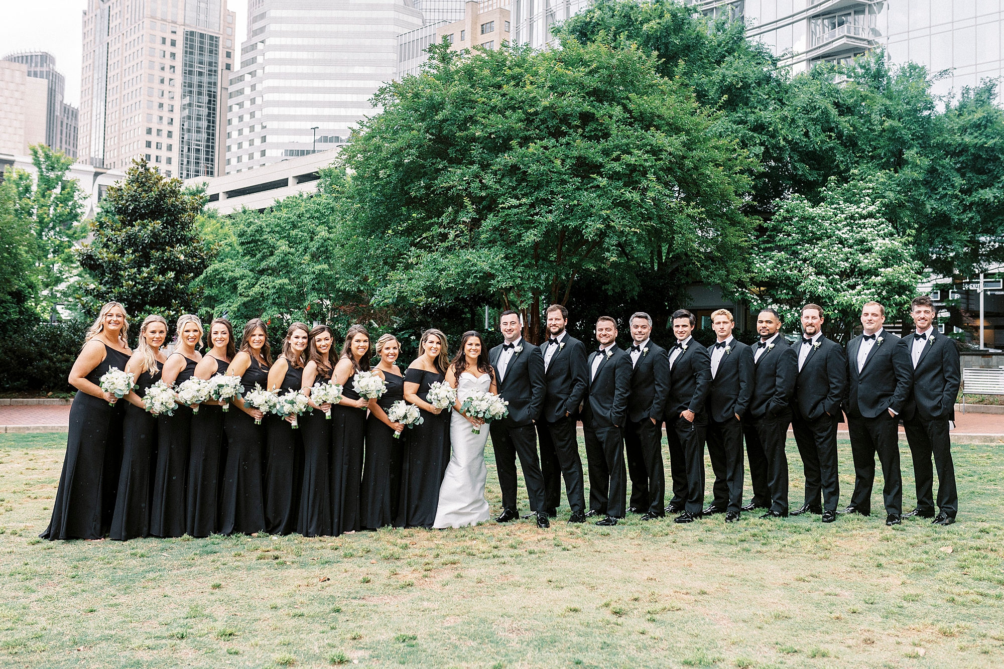 bride and groom stand in park with wedding party in black attire