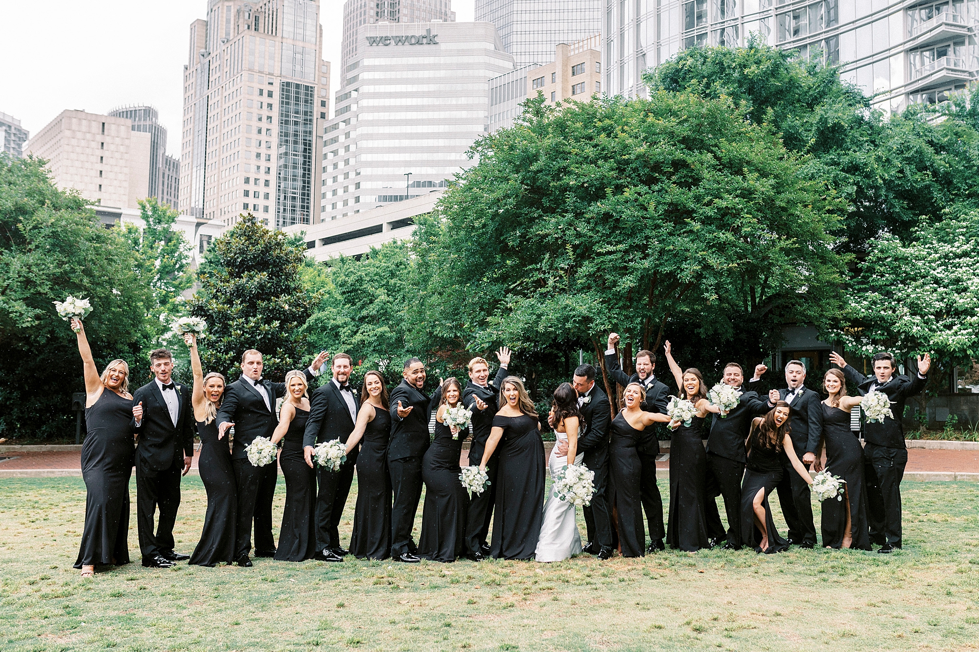 couple kisses while bridal party cheers in Uptown Charlotte park