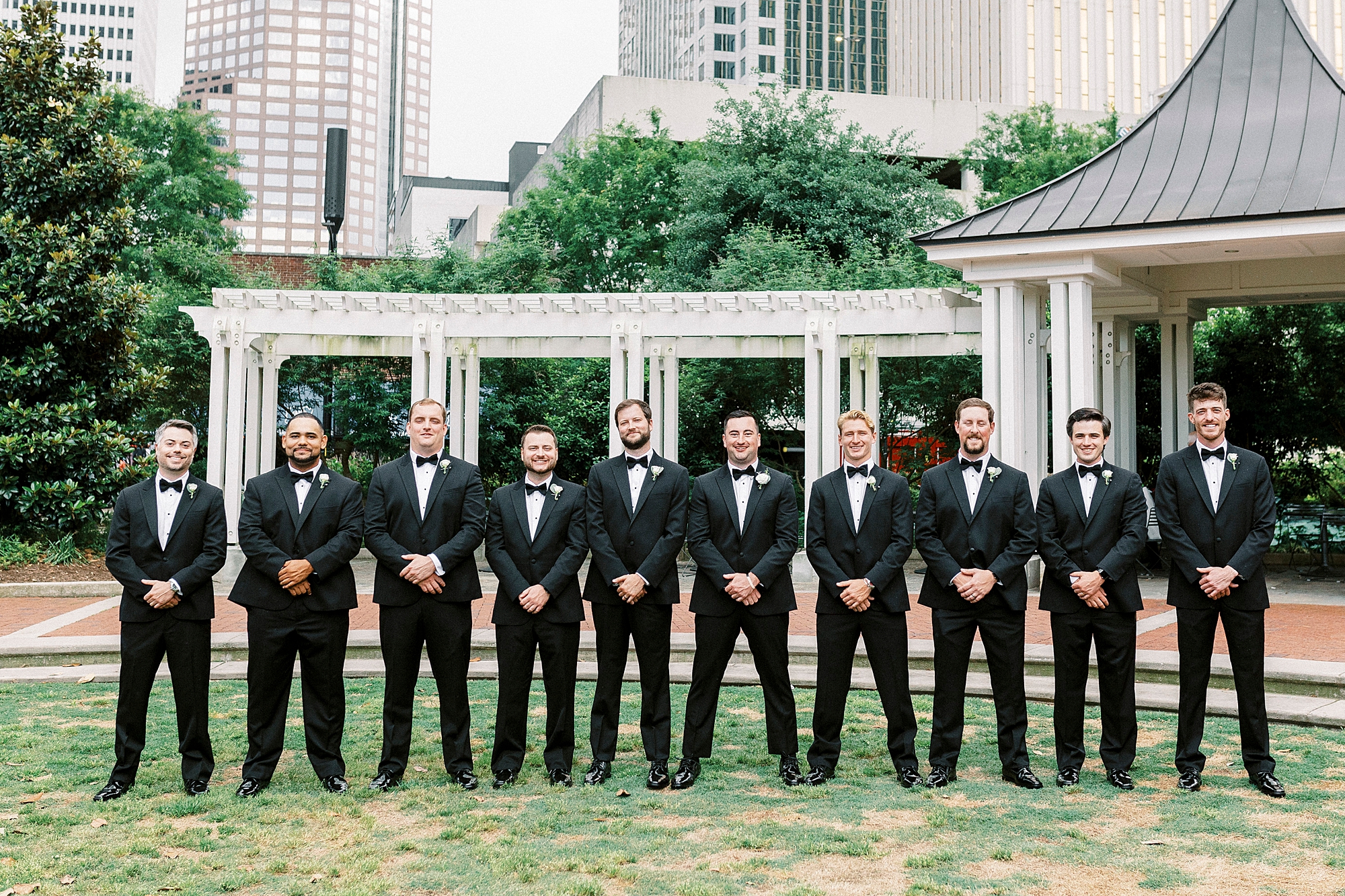 groom stands with groomsmen in tuxes in Charlotte park