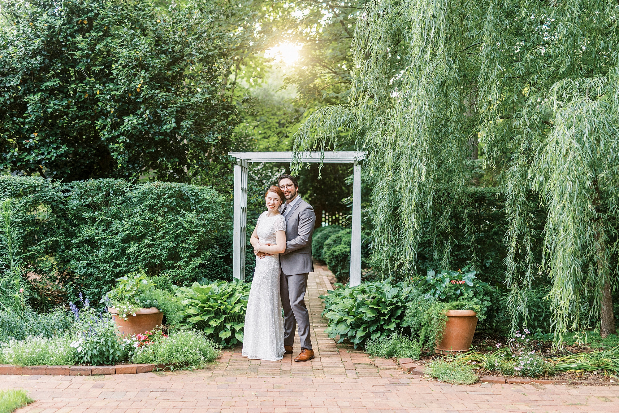 married couple stands under white arbor with man hugging her from behind