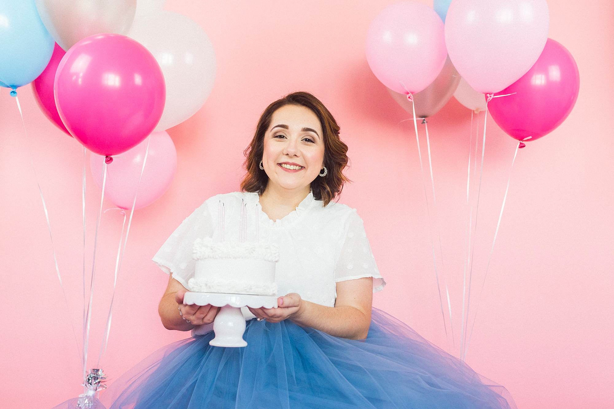 woman sits holding white cake in front of pink backdrop during 30th birthday portraits