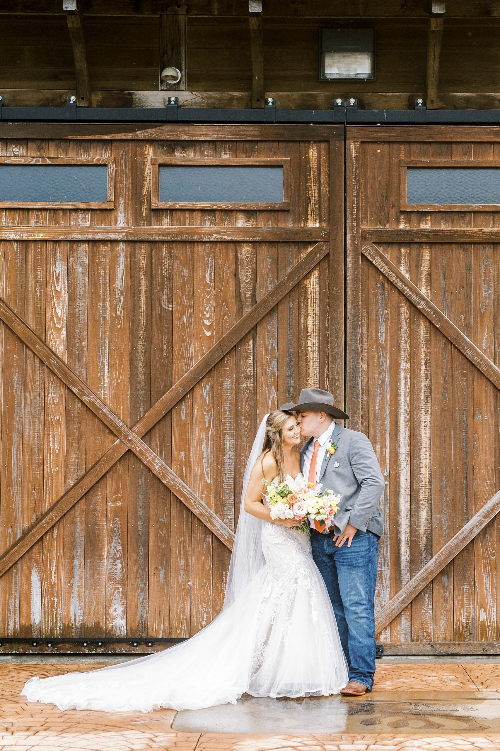 groom in cowboy hat leans to kiss bride's cheek during portraits outside The Stable at Riverview