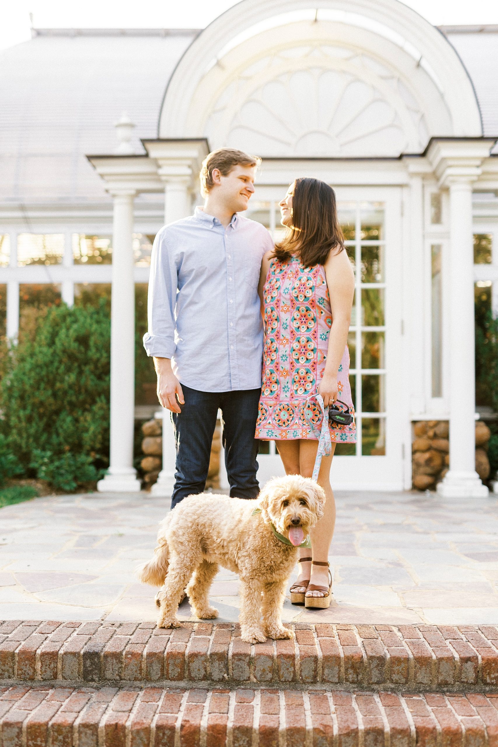engaged couple smiles while dog stands in front of them on steps at Reynolda Gardens