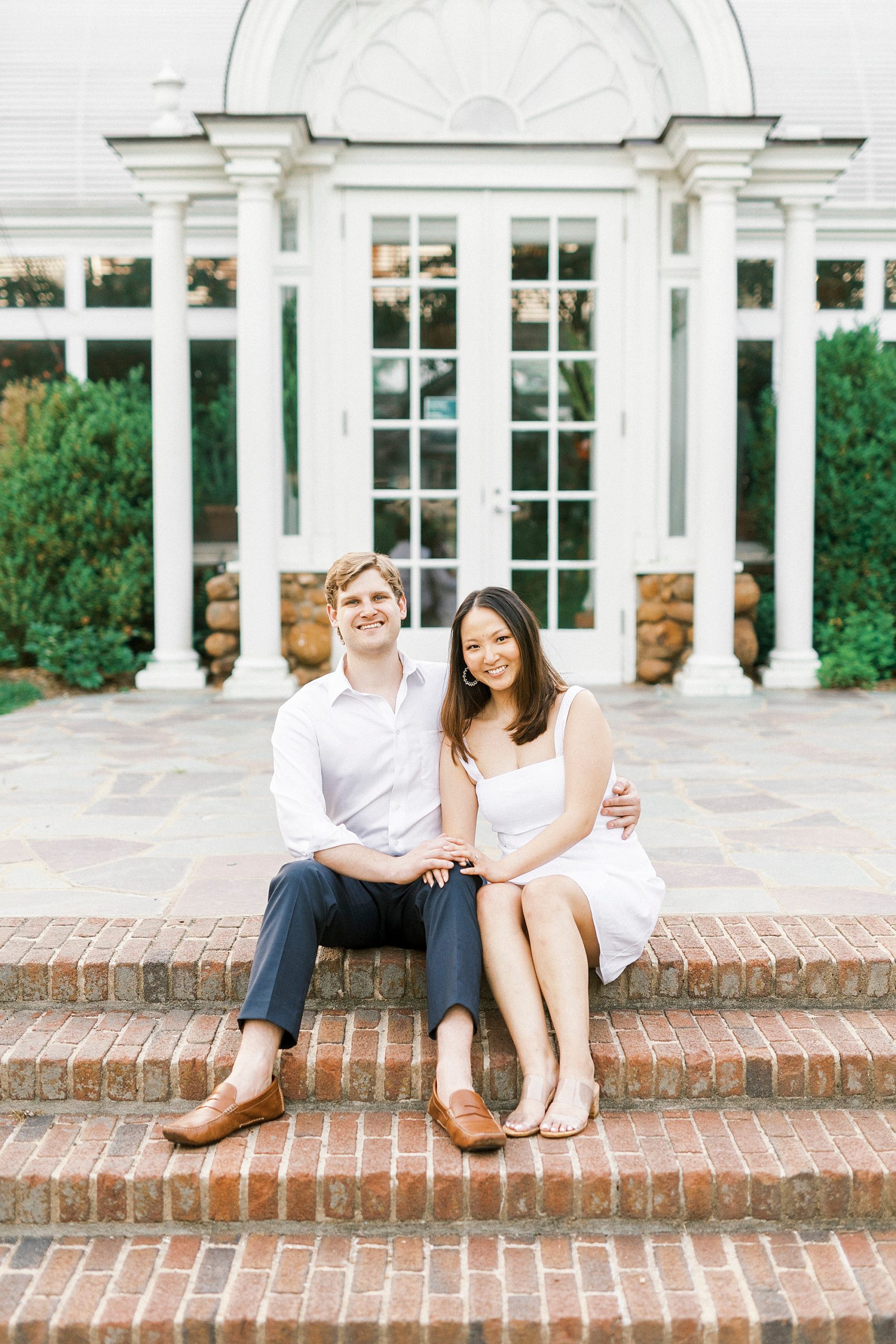 engaged couple laughs sitting on steps in white dress and white button up shirt