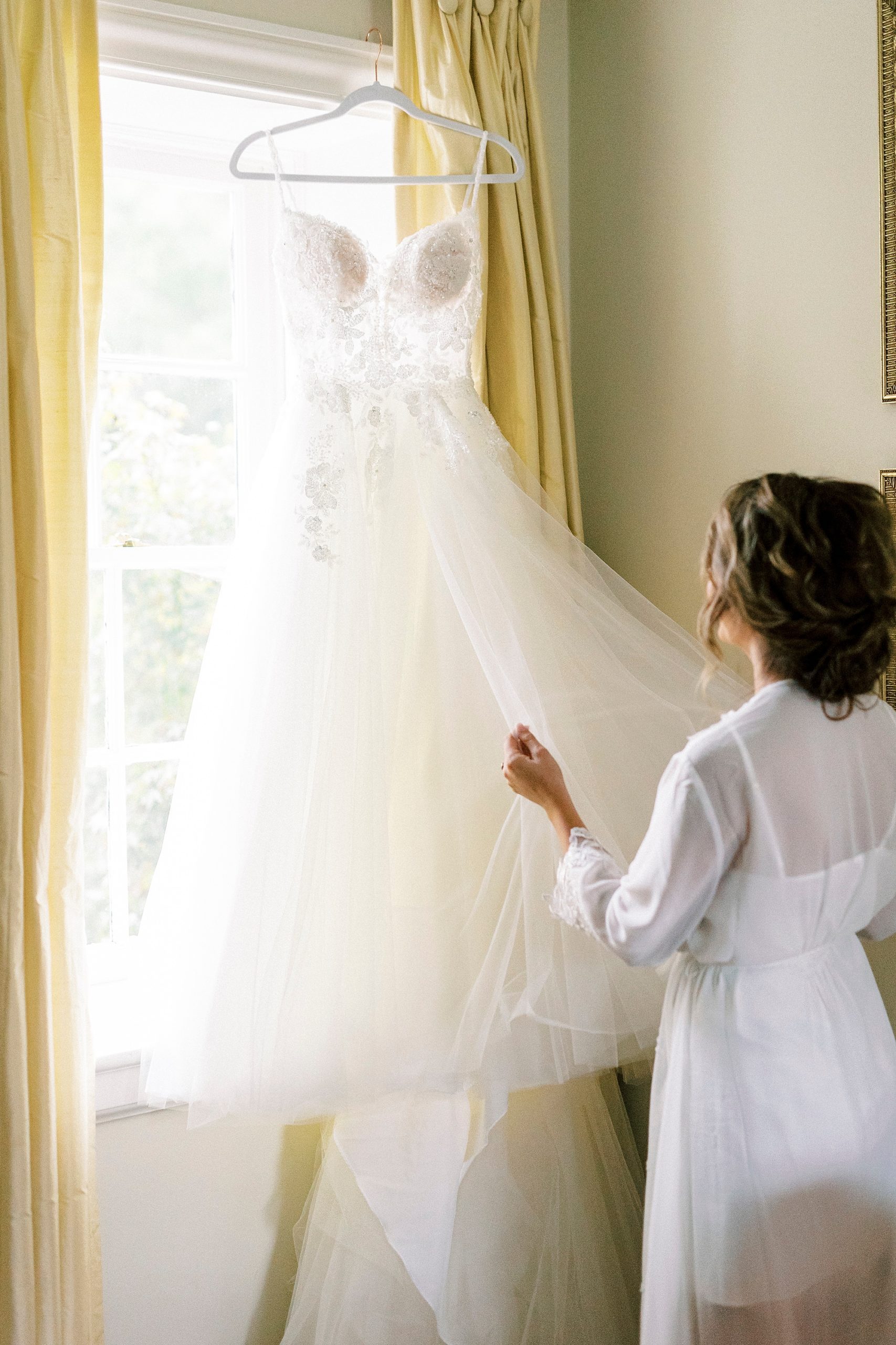 bride looks at wedding dress hanging in window 